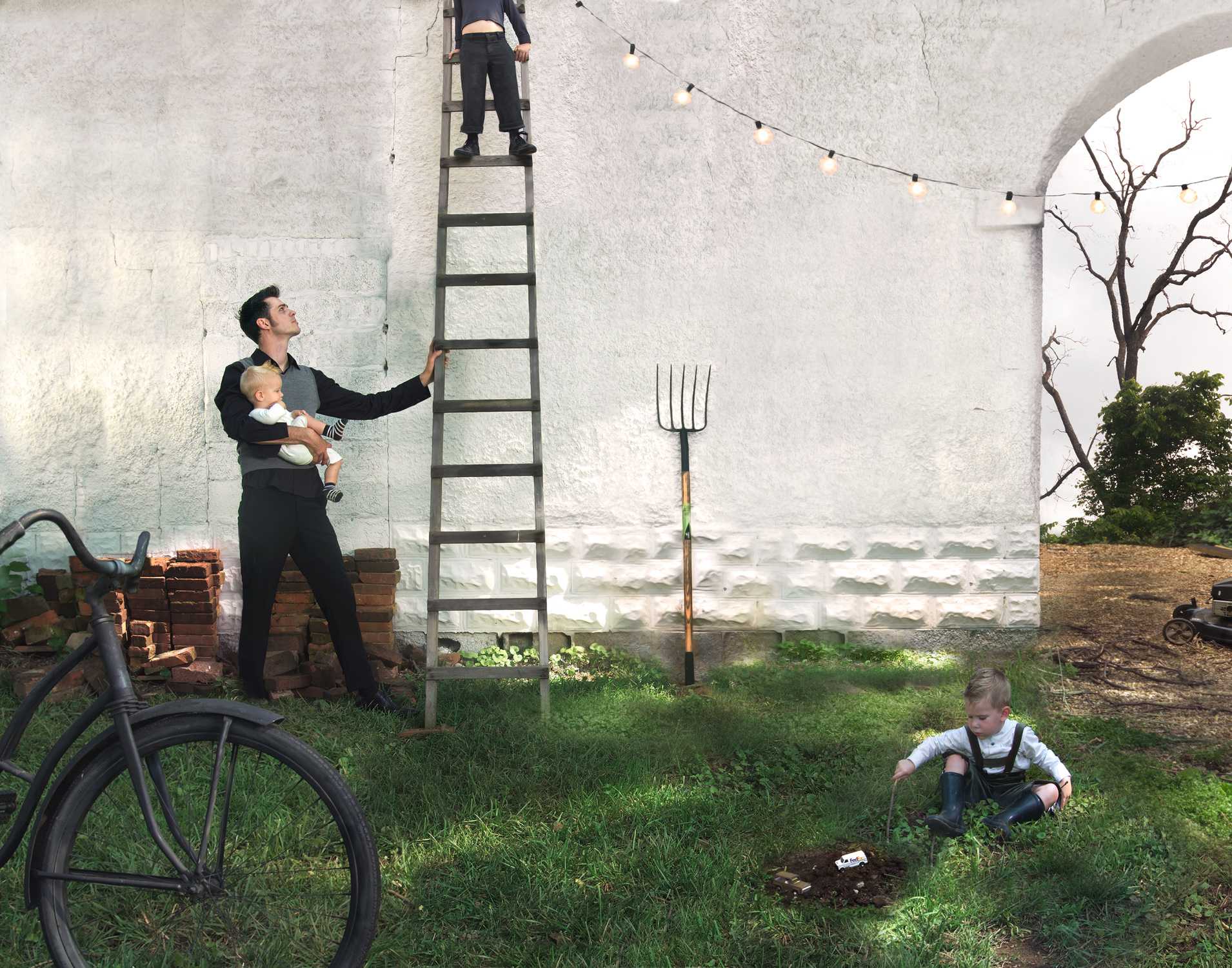 A man in a dark suit holds a small child while steadying a tall wooden ladder against a white concrete wall. A young child in overalls crouches in the grass below, appearing to dig in the soil. A bicycle wheel, gardening fork, bare tree branch, string lights, and scattered bricks are visible in the pastoral scene, creating an absurdist composition that blends domestic and agricultural elements.