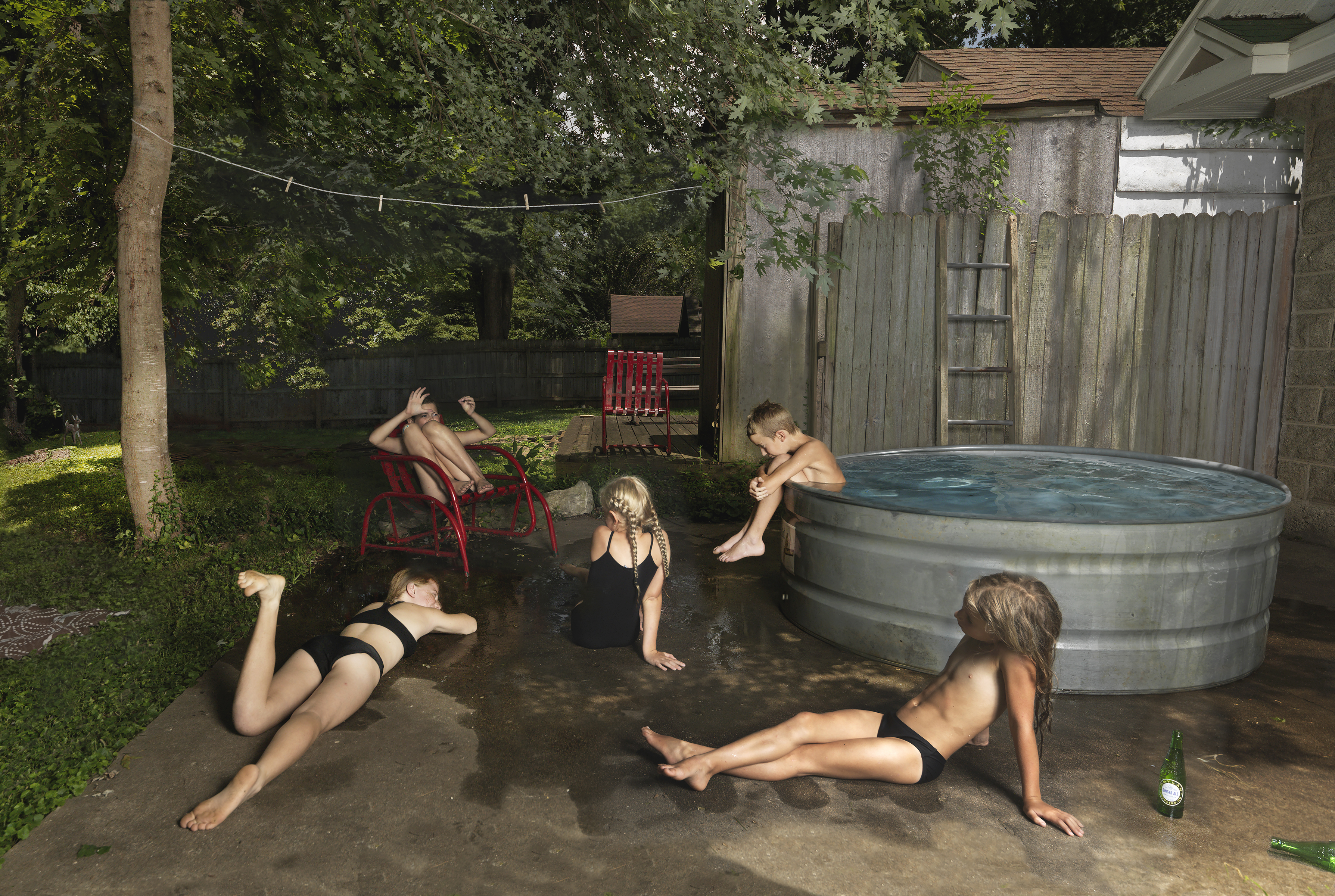 A backyard scene showing six children in swimwear positioned around a large metal stock tank filled with water. Some recline on the concrete ground in various poses of leisure, while others sit in or near the tank and on red Adirondack chairs. Weathered wooden fences, overgrown vegetation, and outbuildings frame the composition, creating a domestic, summery atmosphere rendered in naturalistic color.