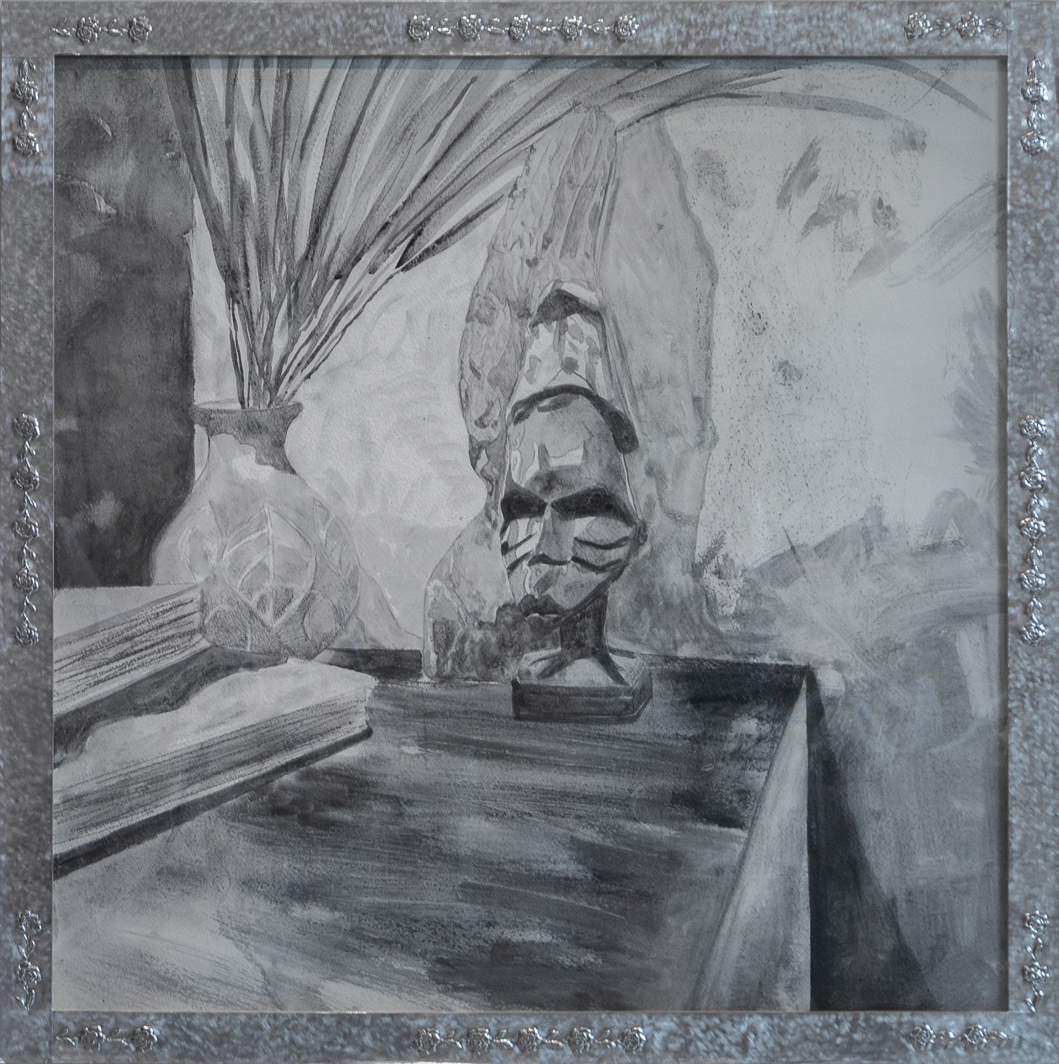 A graphite drawing on gesso depicting a sculptural bust with pronounced facial features and a contemplative expression, positioned on a wooden surface. Dried palm fronds in a decorative vessel are visible to the left, while atmospheric marks and gestural strokes create depth in the background. The work is presented in an ornate silvered frame.