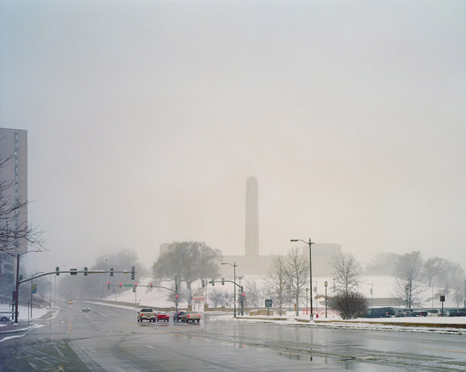 A misty urban street scene with wet pavement reflecting grey skies, featuring a tall cylindrical monument barely visible through heavy fog in the middle distance, bare winter trees lining the street, traffic lights, and parked vehicles along a wide boulevard.