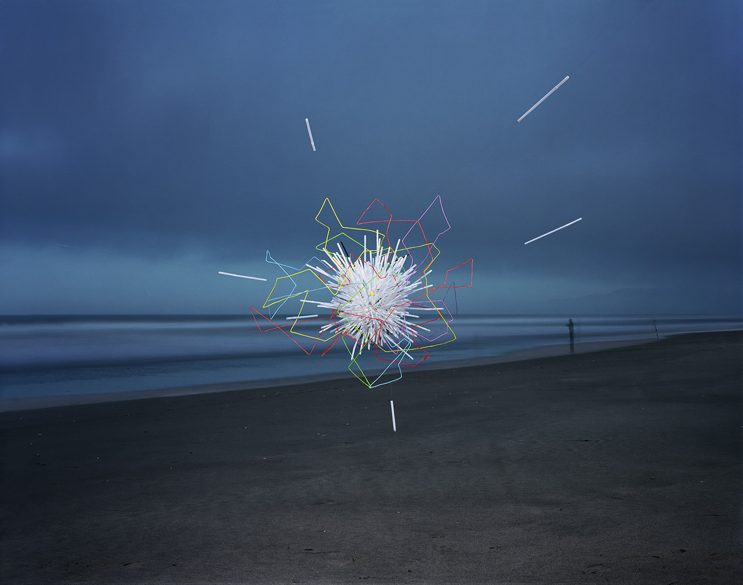 Neon-outlined geometric shapes in yellow, red, pink, and white radiate outward from a bright central burst on a dark beach, with horizontal straws extending across the composition against a moody blue-grey ocean and sky.