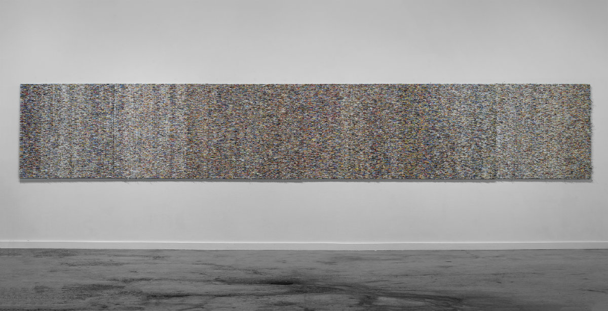 A horizontal rectangular artwork mounted on a white gallery wall composed of densely layered, shredded paper in muted tones of blue, green, brown, and gray, creating an intricate textured surface that reads as an abstract composition from a distance.