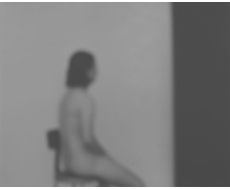 A blurred figure with dark hair sits in profile against a light wall, rendered in soft grayscale tones. The photograph's subtle focus and muted palette create an introspective, contemplative mood.