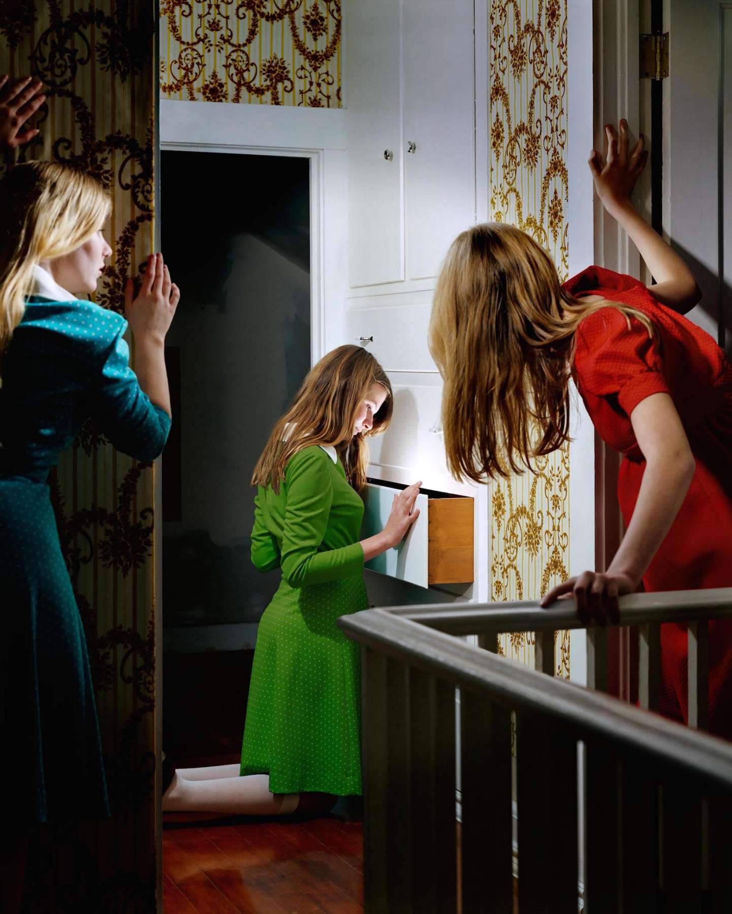Three young girls in brightly colored dresses—turquoise, green, and red—interact with an open white cabinet drawer in an ornate interior with damask-patterned wallpaper. The girl in green leans forward examining the drawer's contents while her companions gesture and look on, their figures bathed in warm, glowing light against the classical architectural backdrop.
