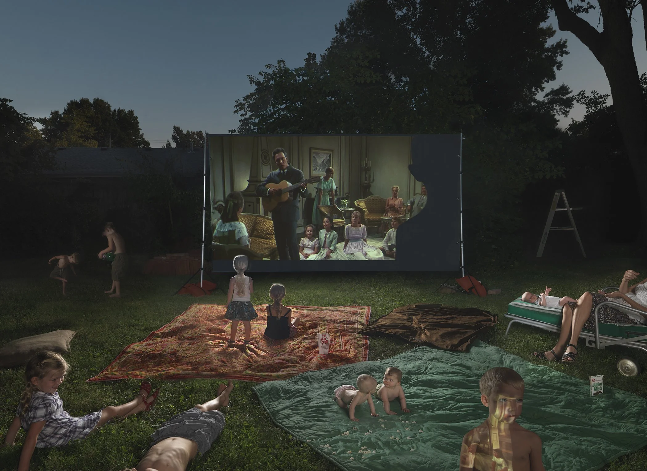 A nighttime backyard scene shows children and adults gathered on a lawn to watch a projected film on an outdoor screen. The projection displays a vintage family scene with people in a mid-century living room, while in the foreground, some children play in an inflatable pool, others lounge on blankets and chairs, and figures rest on the grass, all illuminated by the glow of the large screen against a darkened tree line.