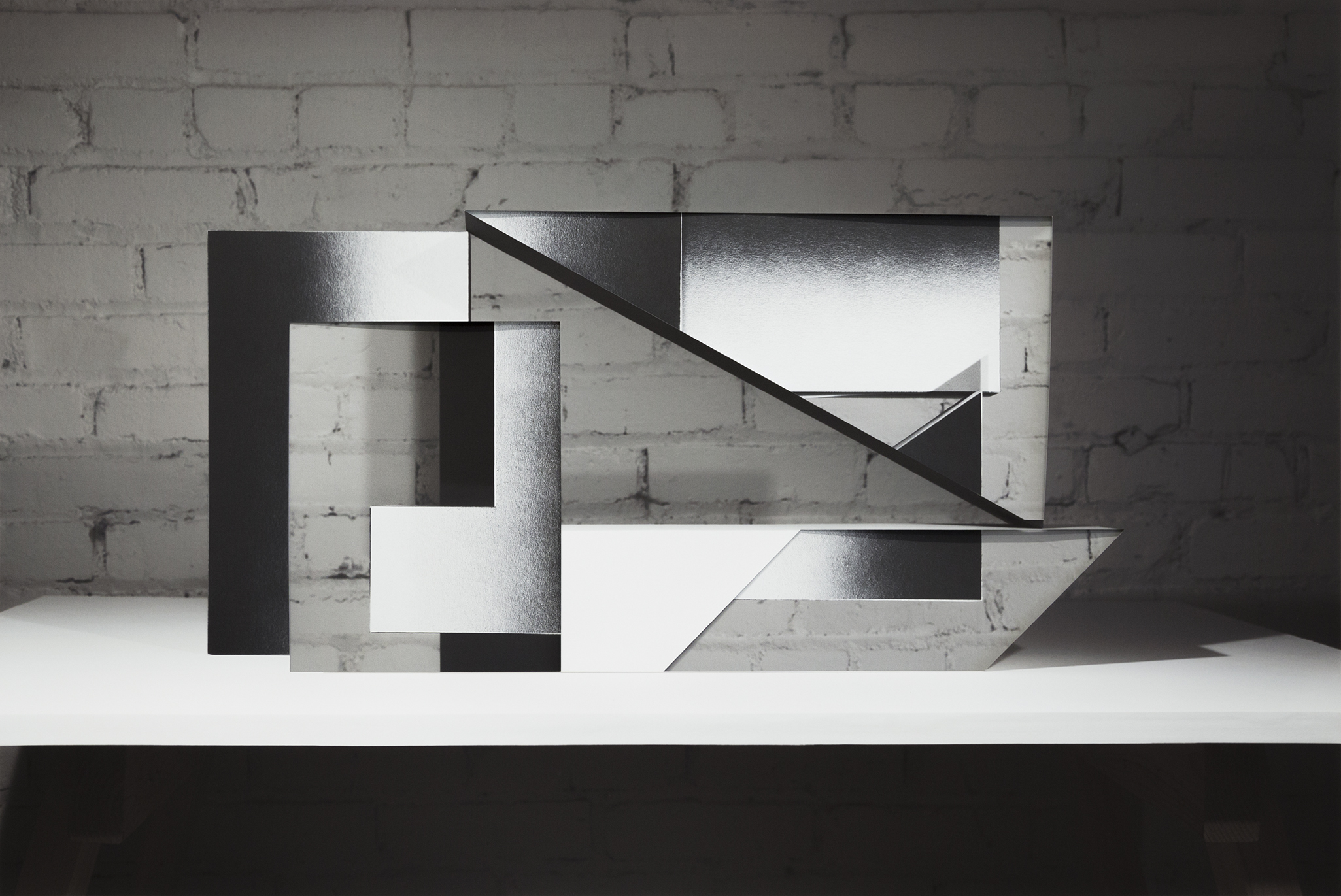 An abstract sculptural composition of layered black and white rectangular forms arranged on a white platform against a white brick wall. The overlapping planes create sharp angles and diagonal lines, with contrasting matte and reflective surfaces that cast distinct shadows.
