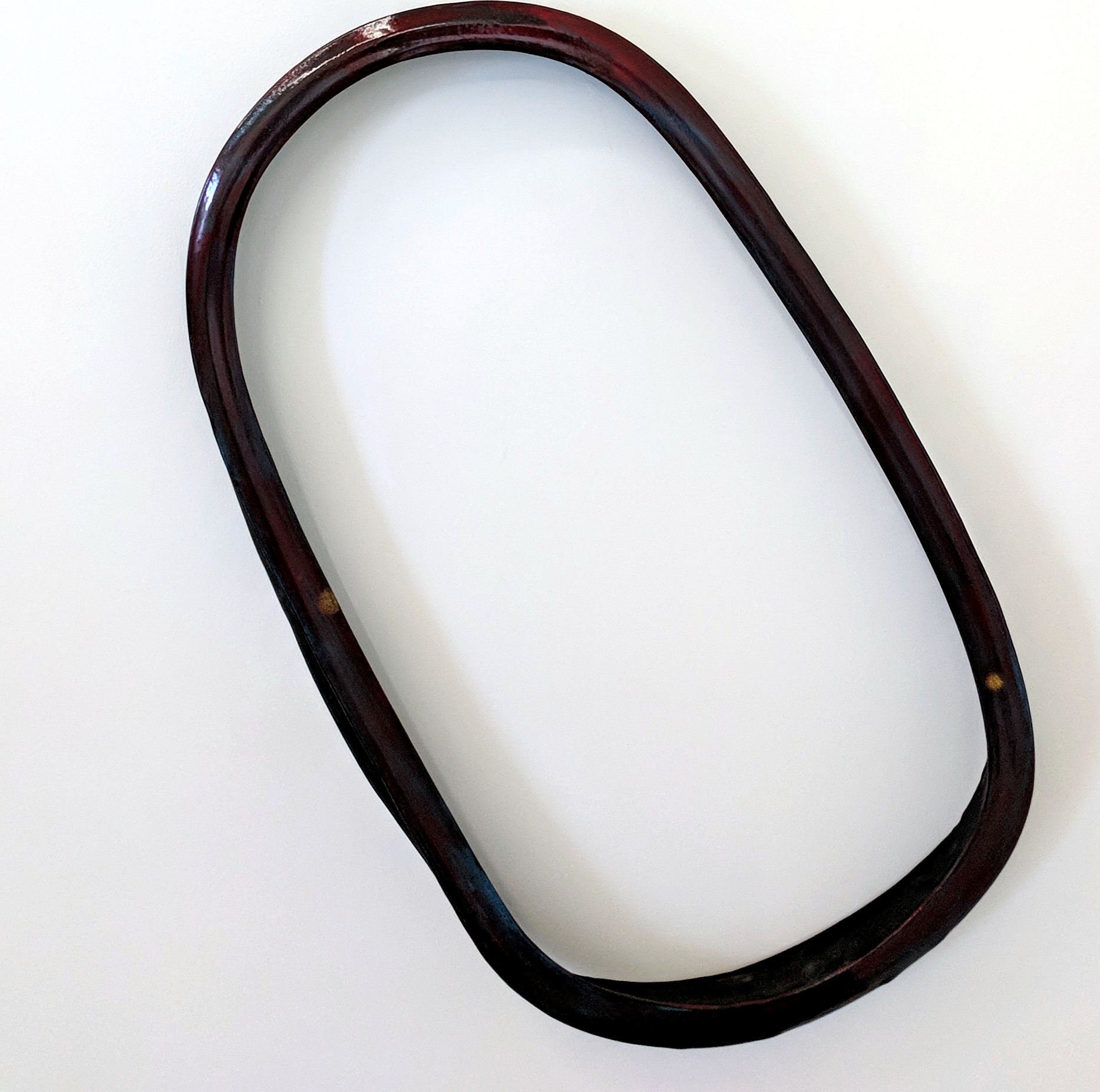 A dark reddish-brown ceramic ring with a smooth, curved form sits against a white background. The sculptural object has an organic, minimalist aesthetic with subtle variations in tone and a polished surface.