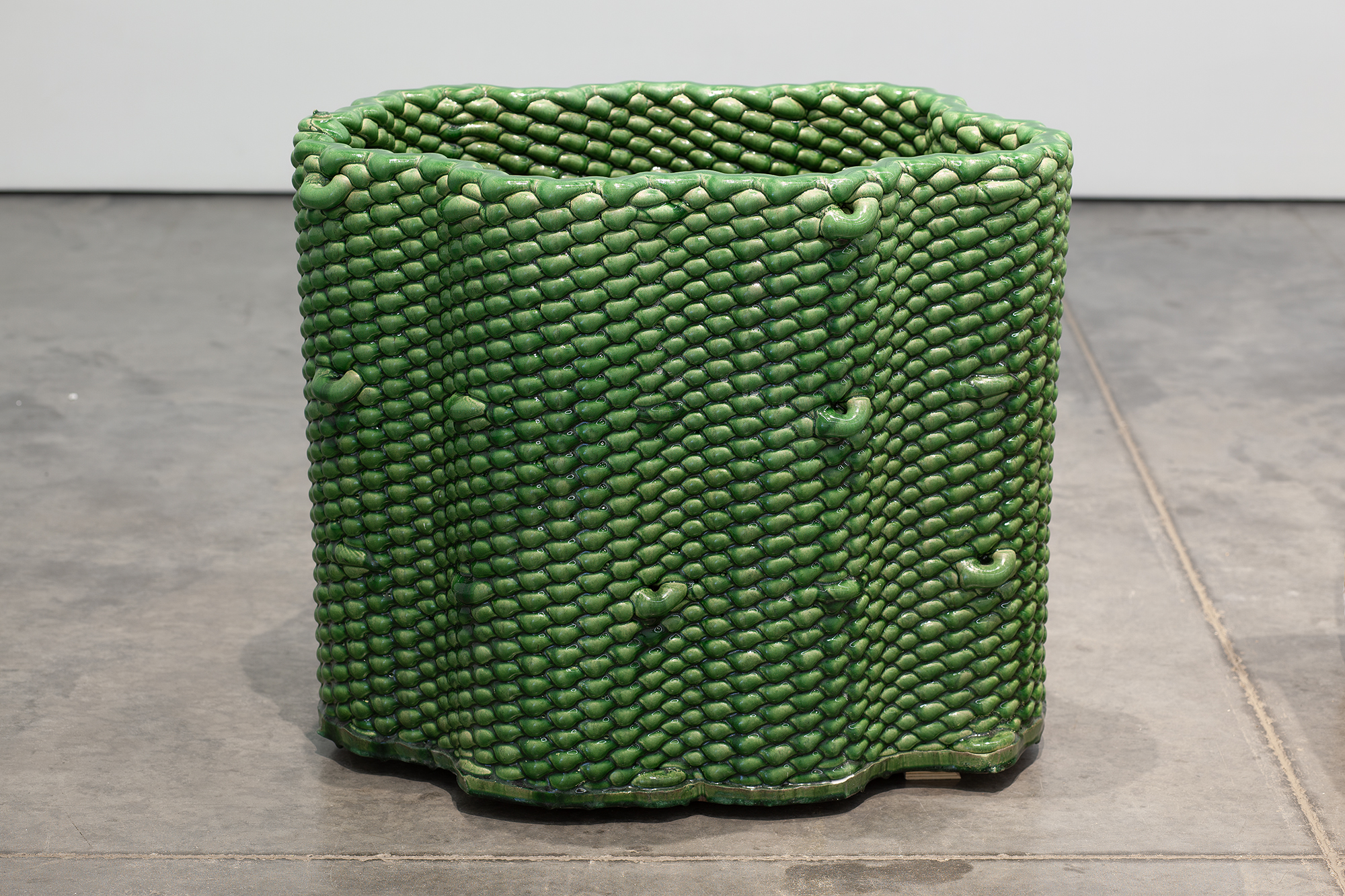 A cylindrical vessel with a textured surface composed of densely packed small rounded forms in green glaze, displayed on a concrete floor against a white wall.