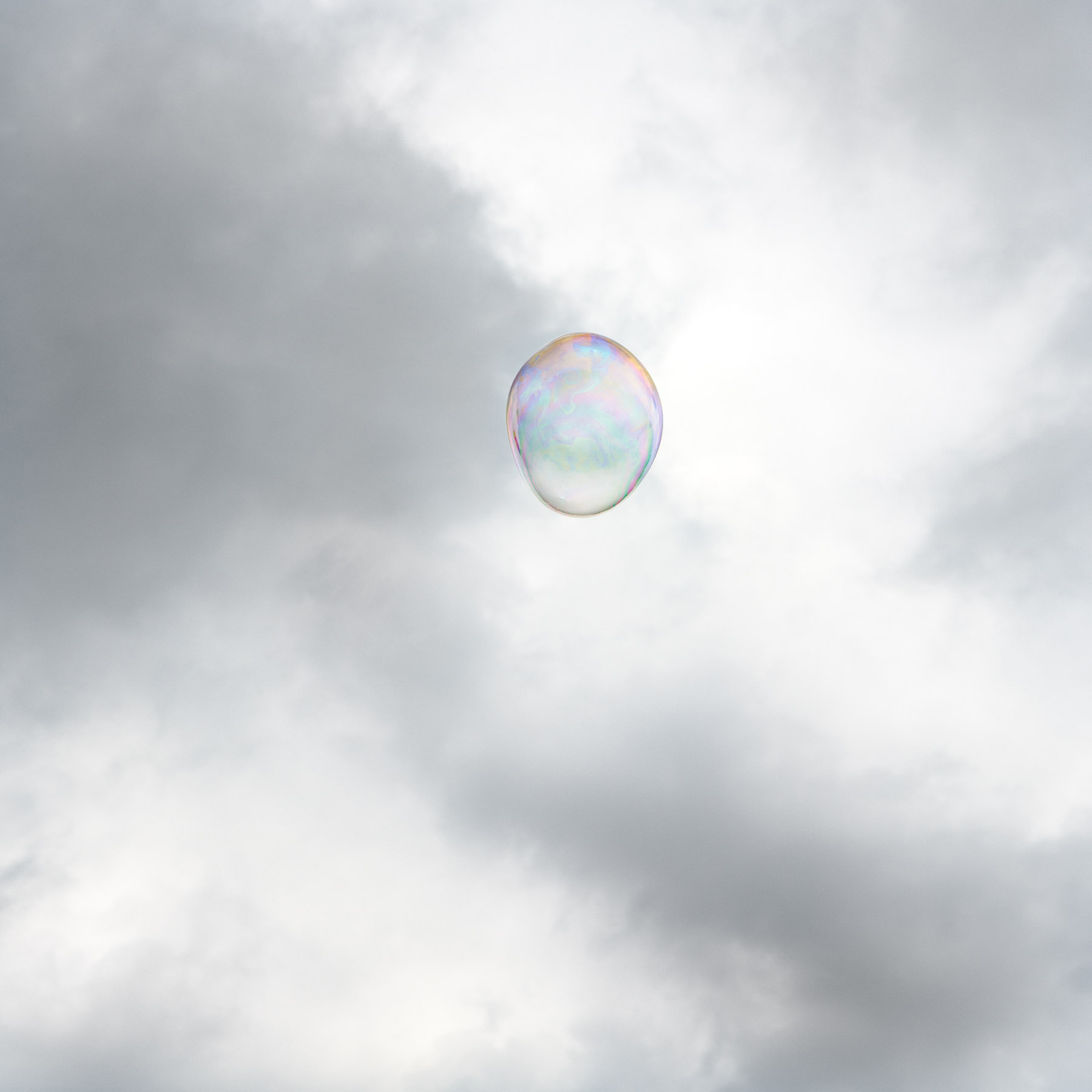 A single iridescent soap bubble floats against a cloudy gray and white sky, its translucent surface reflecting soft pastel hues of pink, blue, and green.
