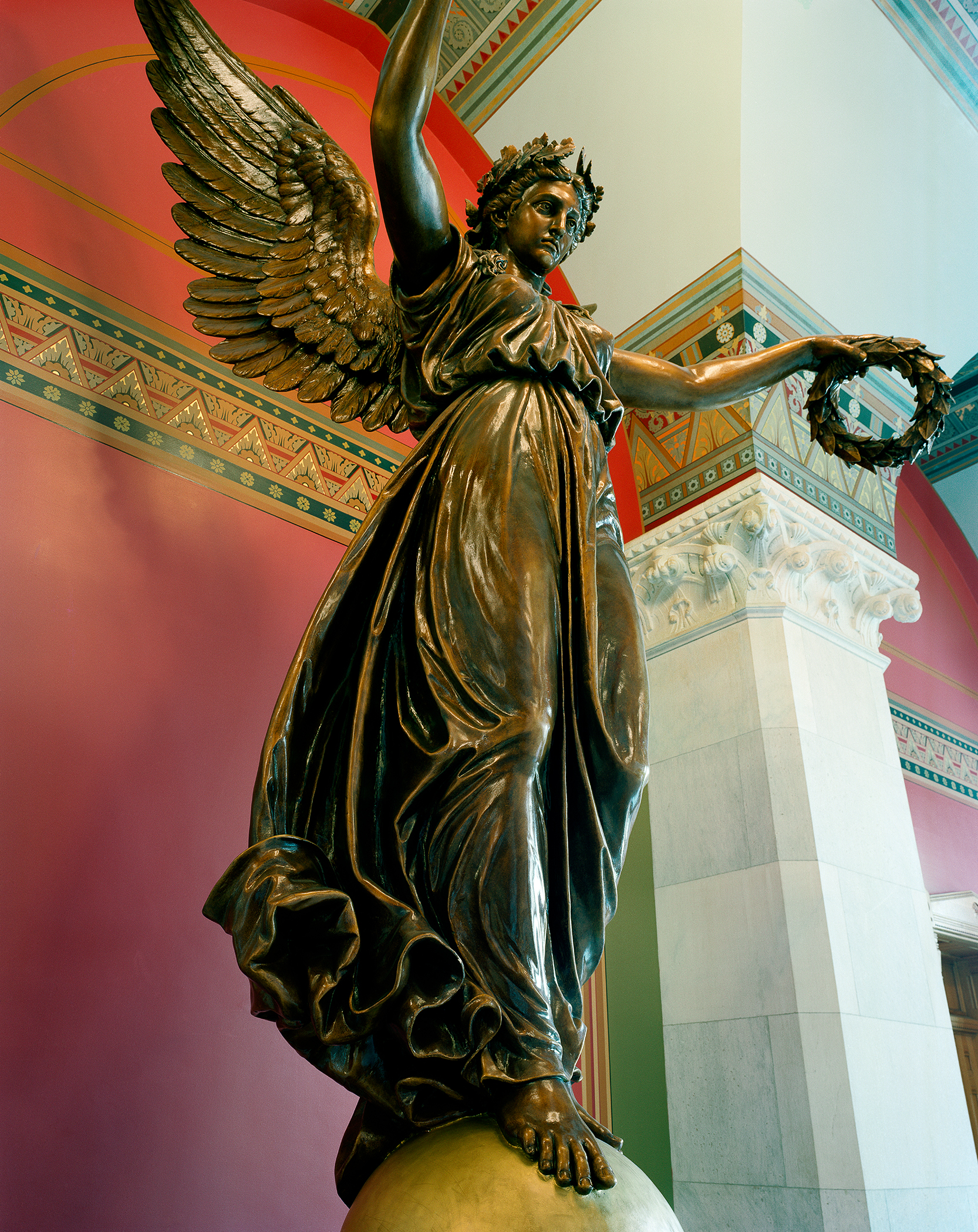 Bronze winged angel statue with laurel wreath, raising one arm upward while standing on a draped pedestal, photographed against a vibrant red wall with ornamental green and gold architectural detailing visible in the background.