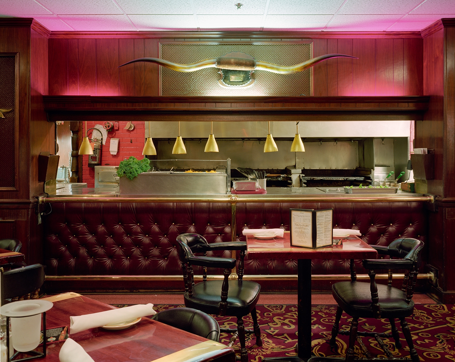 A luxurious restaurant interior dominated by deep burgundy leather banquette seating and rich wood paneling. The open kitchen features stainless steel countertops and brass pendant lights, while a large taxidermied longhorn with golden horns is mounted above the bar, creating a striking focal point against the pink-lit back wall. Ornate red and gold patterned carpeting and black leather chairs complete the upscale, retro-inspired aesthetic.