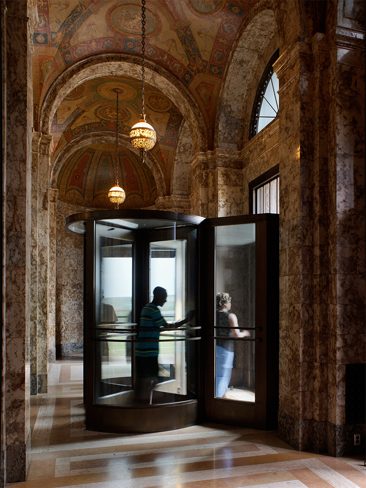 A curved black glass and metal structure stands in an ornate corridor with barrel-vaulted ceilings decorated in faded frescoes and geometric patterns. Two figures are visible through the glass doors against bright exterior light, while ornate brass pendant lights hang from the ceiling above the polished terrazzo floor.