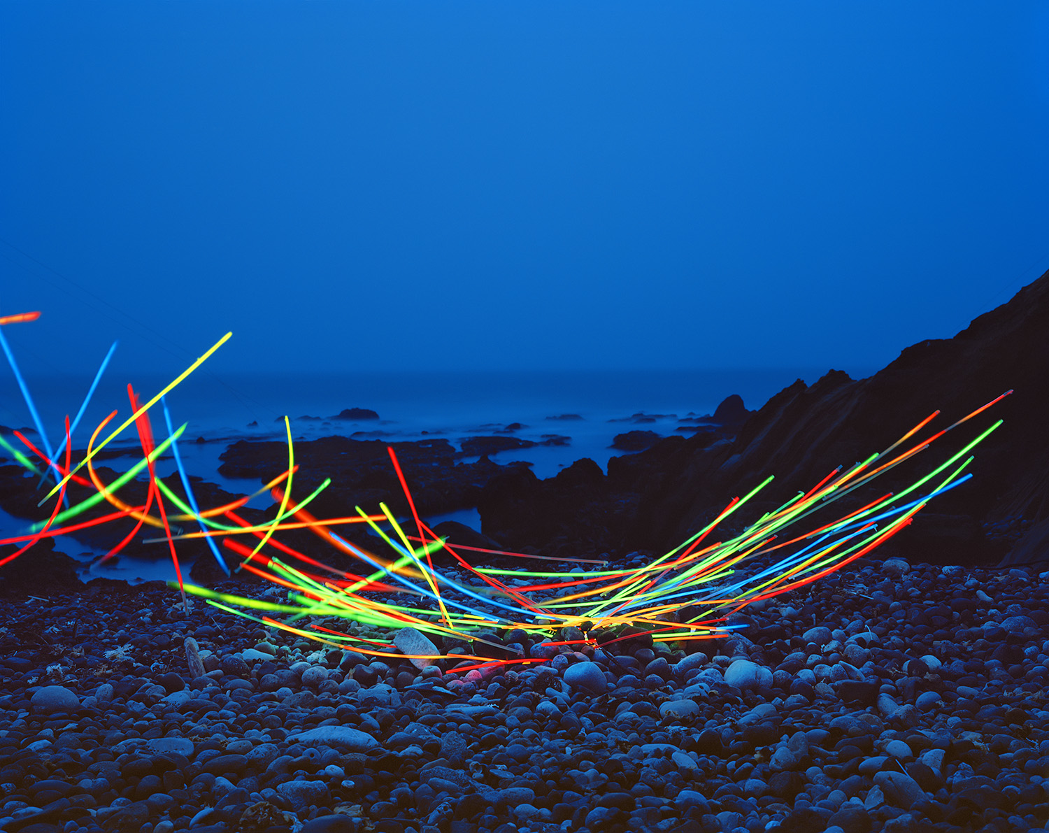 Neon glow sticks arranged in tangled, radiating patterns across a rocky beach at dusk, their bright red, yellow, green, and blue lines creating dynamic linear forms against the darkened landscape and twilight sky.