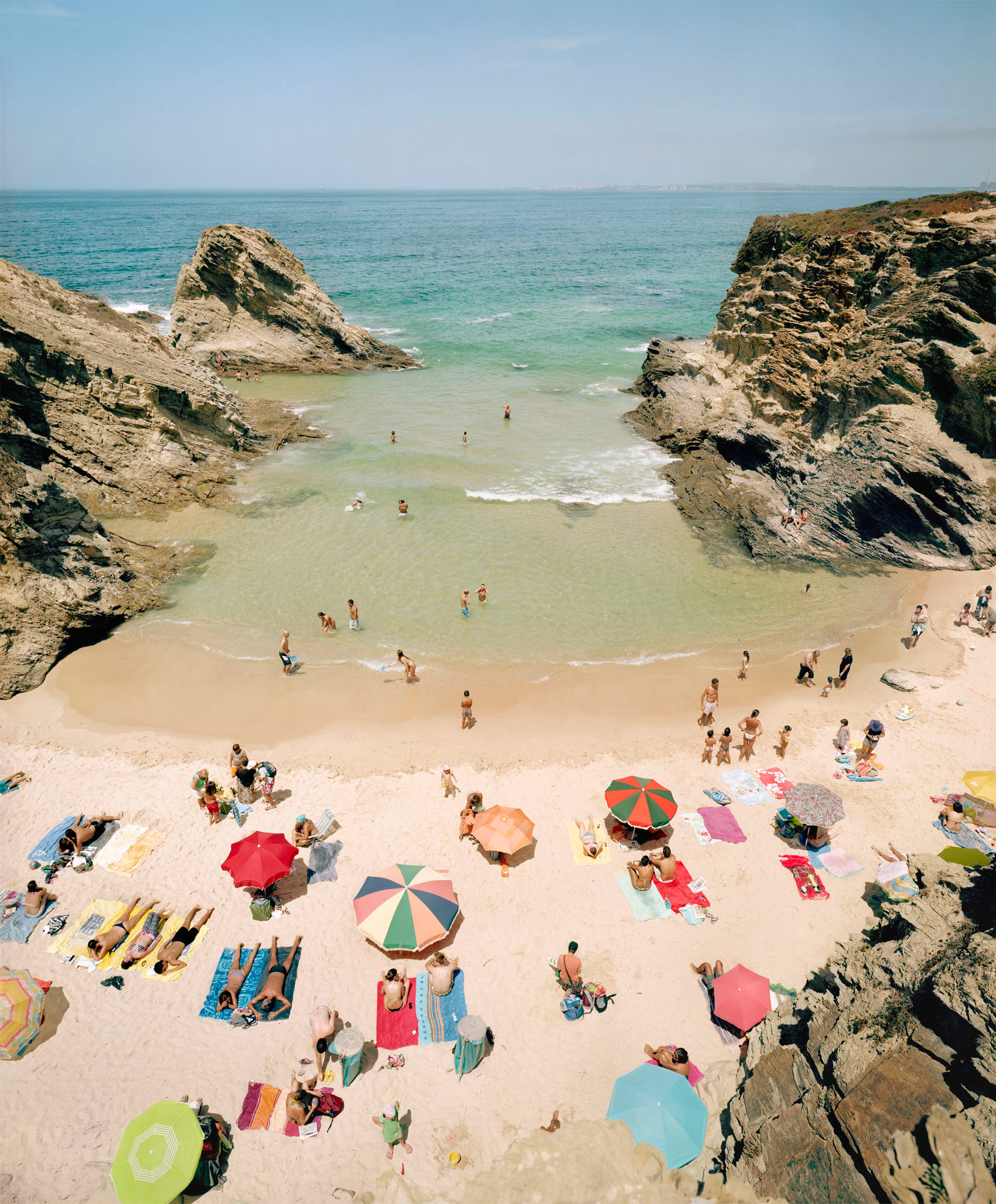 Aerial view of a crowded sandy cove bordered by dramatic rocky outcrops, with beachgoers scattered across the sand beneath colorful umbrellas and on towels, while others wade in the turquoise and pale green waters below.