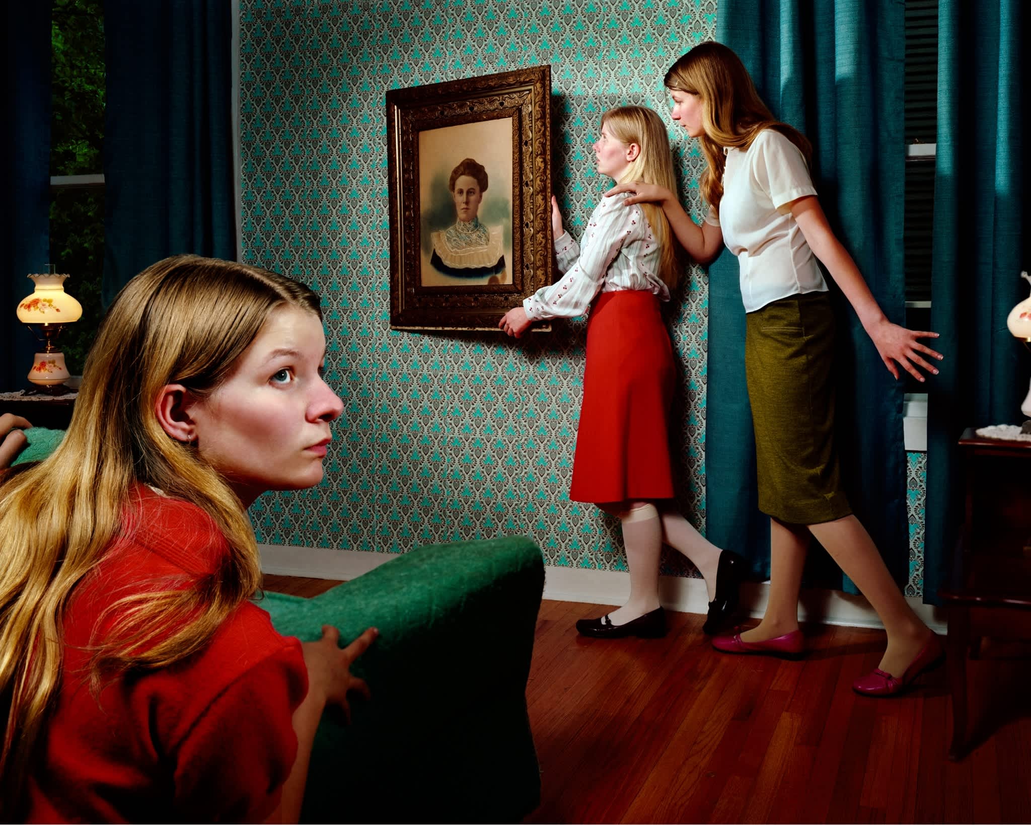 A staged interior scene with turquoise patterned wallpaper and teal curtains. Three figures in mid-20th century clothing examine a framed portrait on the wall, while a fourth figure in a red coat sits in the foreground on a green chair, gazing away from the group. Warm lamps and wooden furniture complete the richly colored domestic setting.