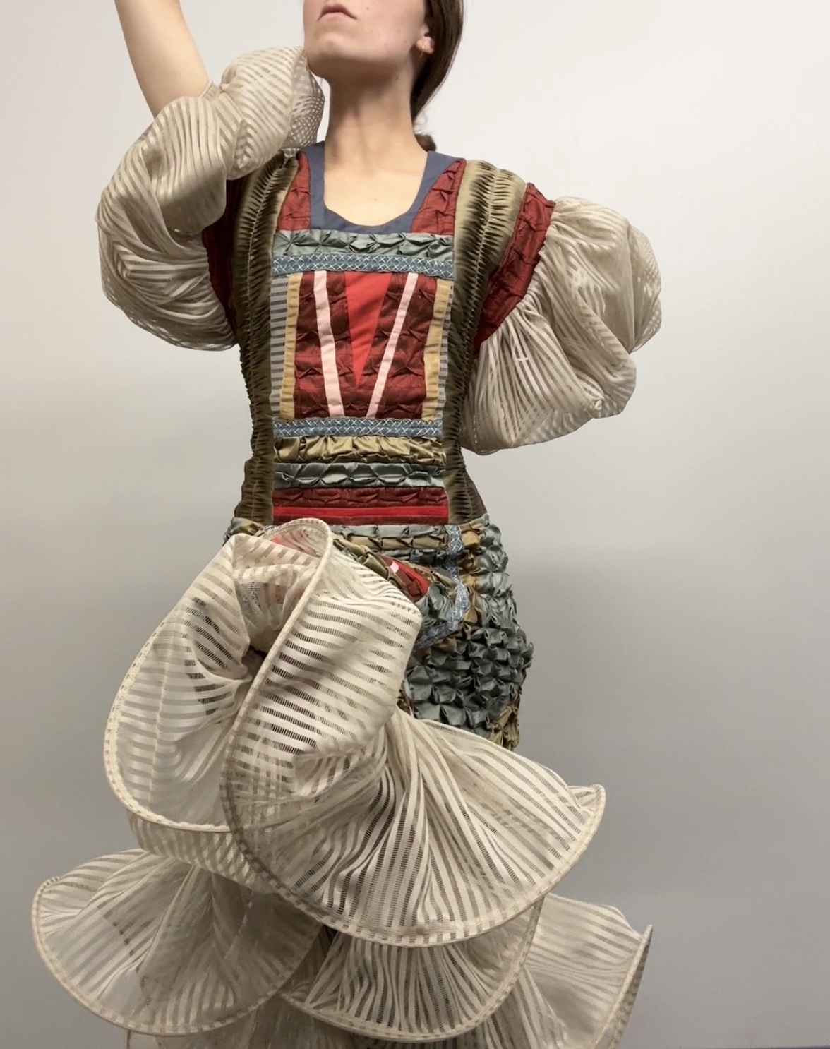 A dress form wearing a sculptural garment composed of pleated cream-colored taffeta forming dramatic ruffled layers at the skirt, paired with a structured bodice featuring a patchwork design in red, tan, and muted green tones with geometric embroidered patterns and decorative striping. The garment is accessorized with voluminous pleated puffed sleeves in matching cream taffeta.