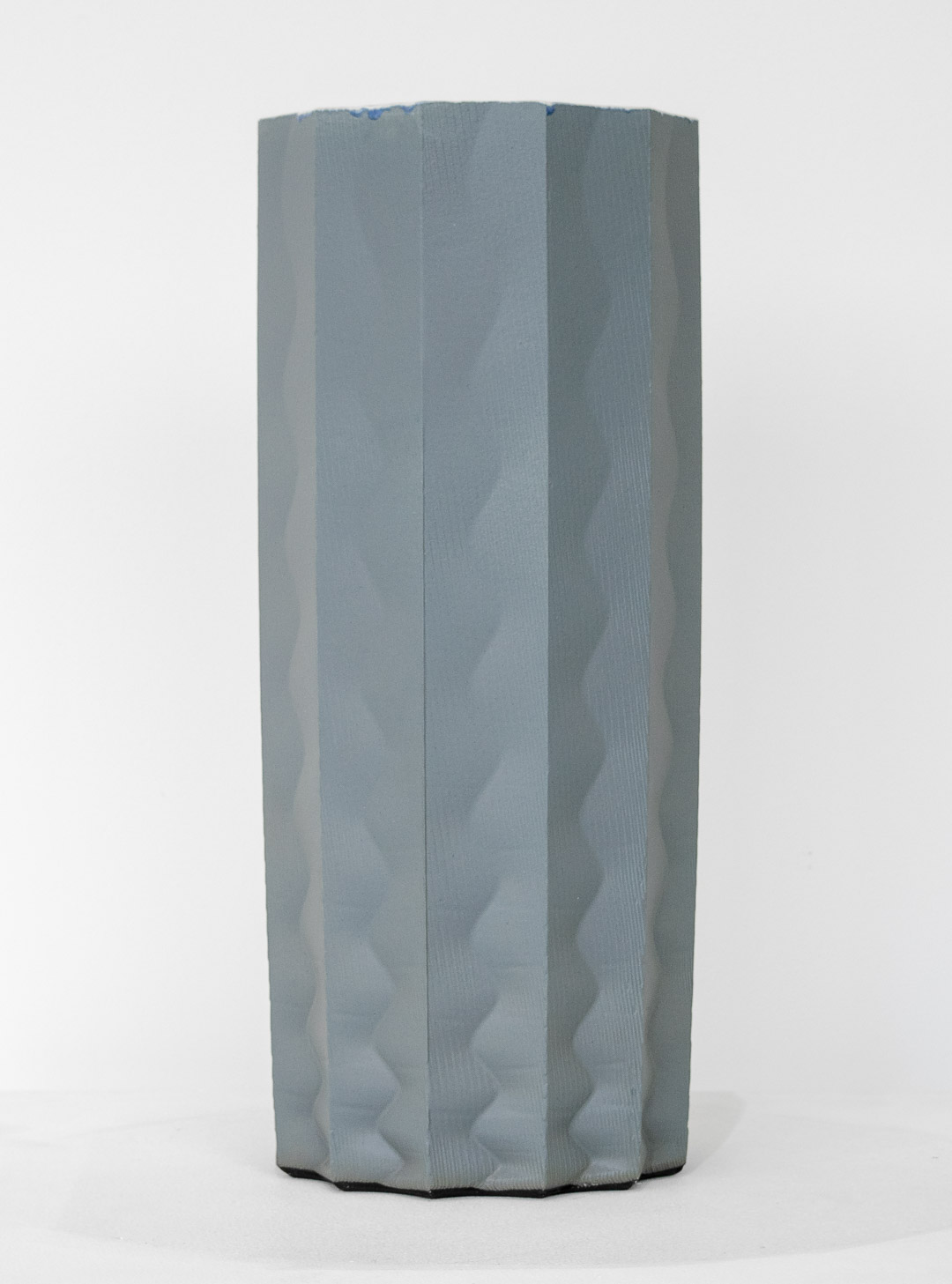 A tall cylindrical porcelain form with vertical ridged panels in soft gray and white tones, featuring subtle wave-like undulations across its surface. The sculpture stands against a neutral background, its organic, fluted structure creating areas of shadow and depth.