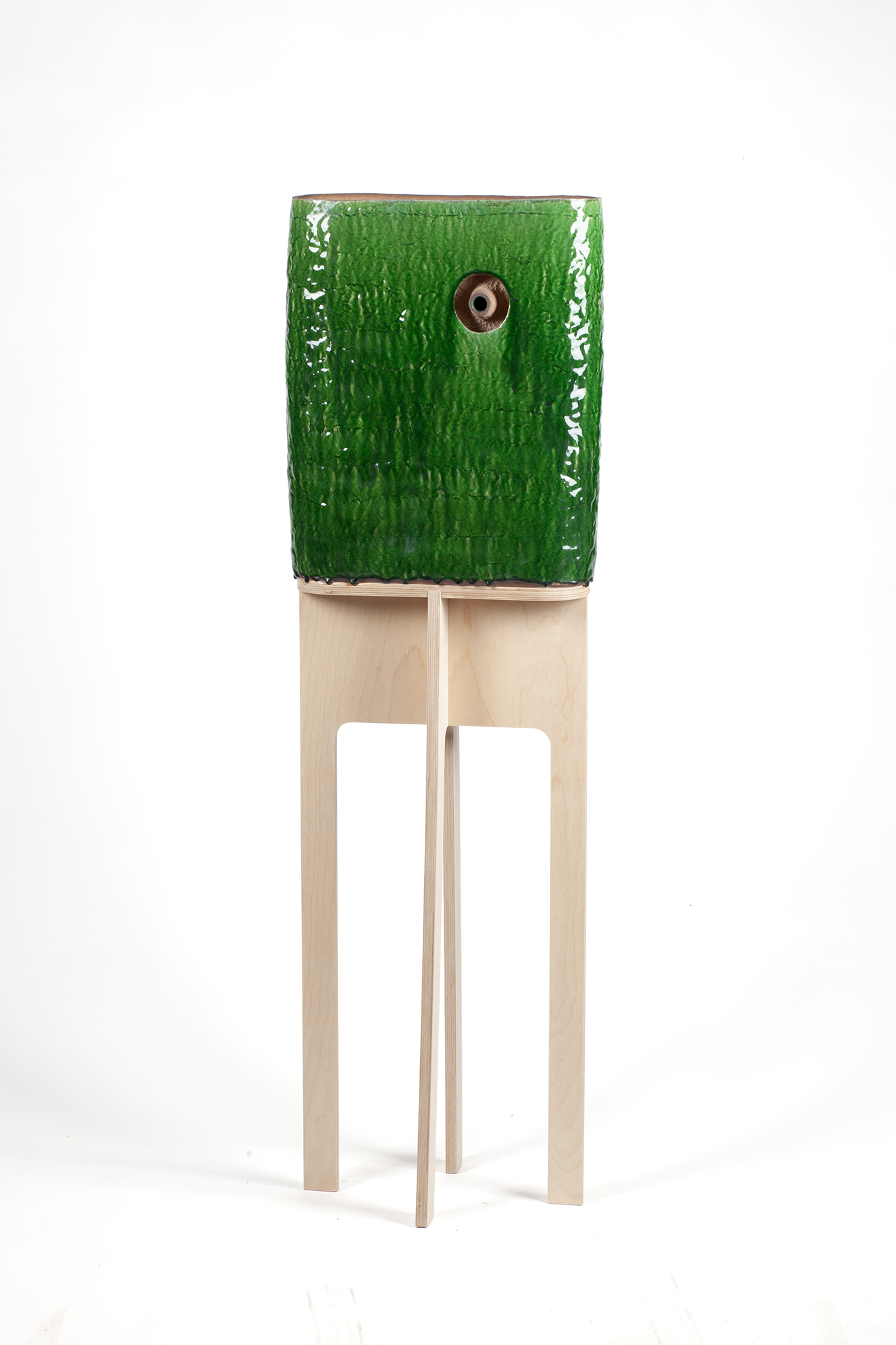 A glazed ceramic head with a bright green textured surface sits atop a natural wood stand with four splayed legs. A single circular opening serves as the gaze, creating an anthropomorphic form that appears to observe its surroundings.