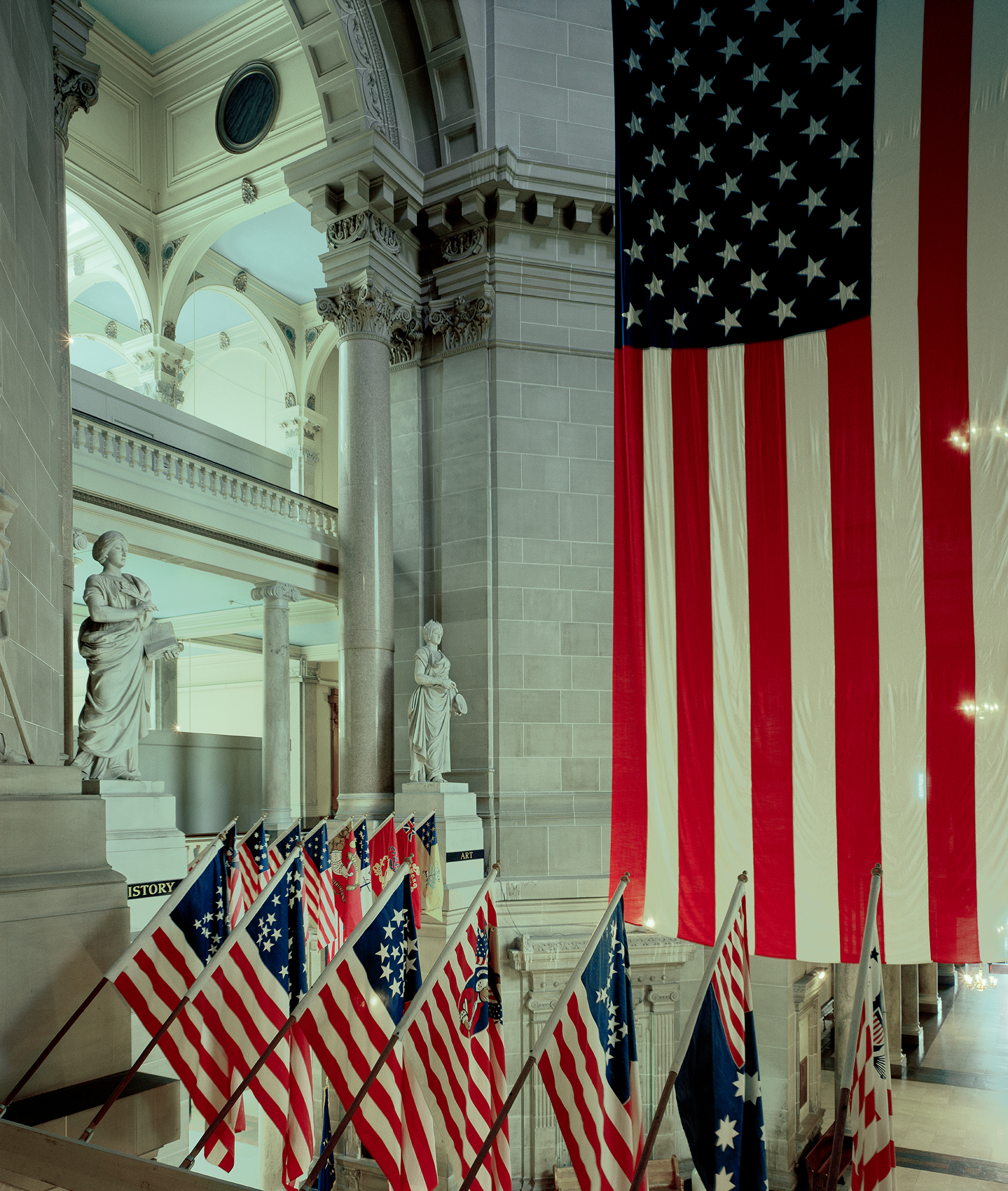 American flags in various historical designs are displayed throughout a classical neoclassical interior with ornate columns, arches, and marble statuary, creating a composition that juxtaposes patriotic symbolism with architectural grandeur.