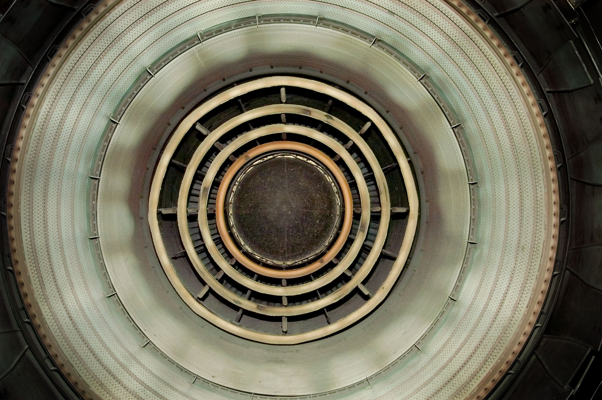Concentric circles of varying widths and textures recede toward a dark center, creating a tunnel-like perspective. The rings transition from pale green and cream tones in the outer layers to darker blacks and browns toward the core, with visible mechanical elements and rivets suggesting industrial machinery or mechanical systems.