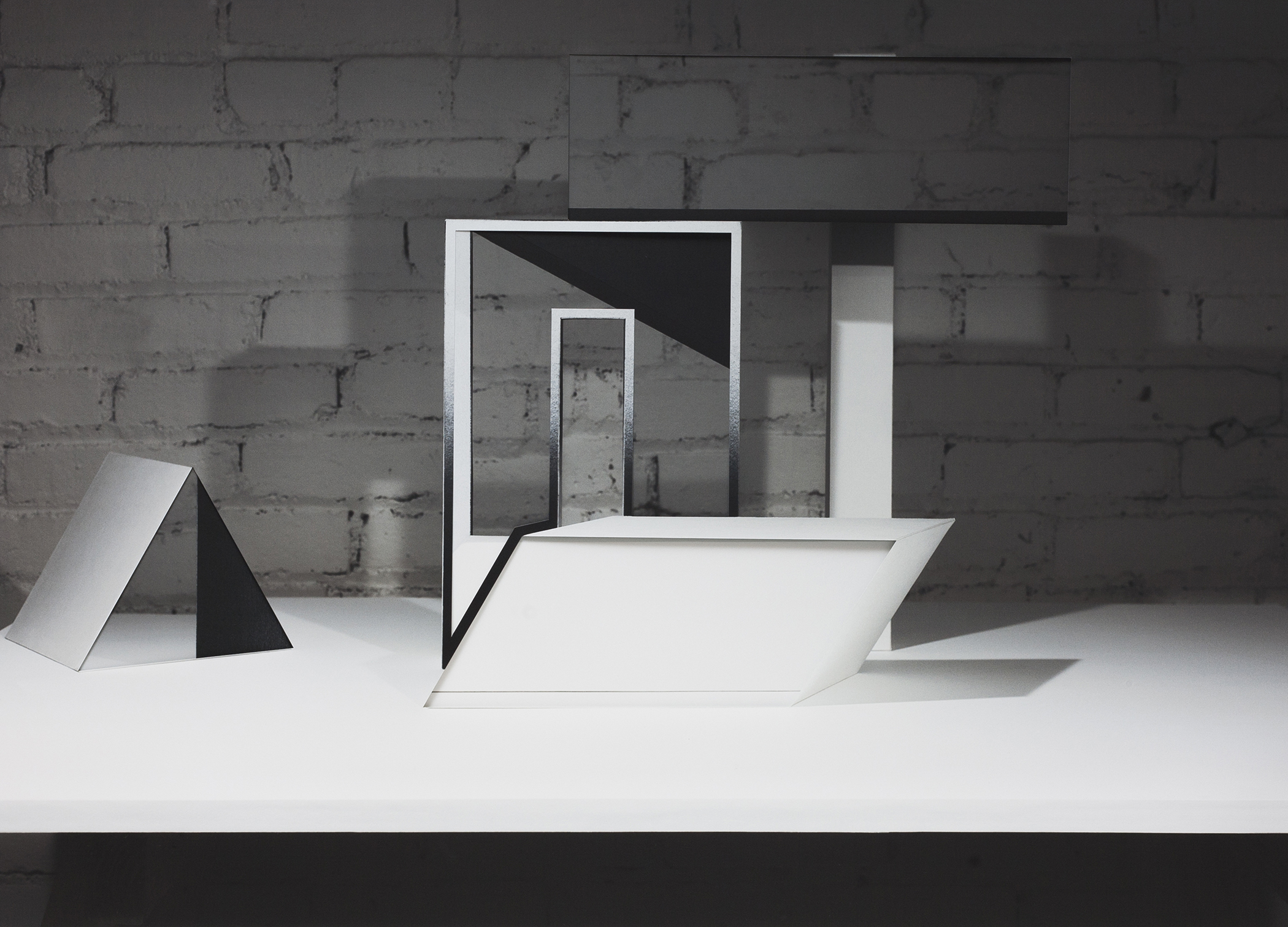 White and black geometric paper forms arranged on a white platform against a weathered brick wall. The sculptural composition features angular planes, rectangular frames, and triangular shapes in varying scales, creating an abstract architectural study in monochromatic tones.