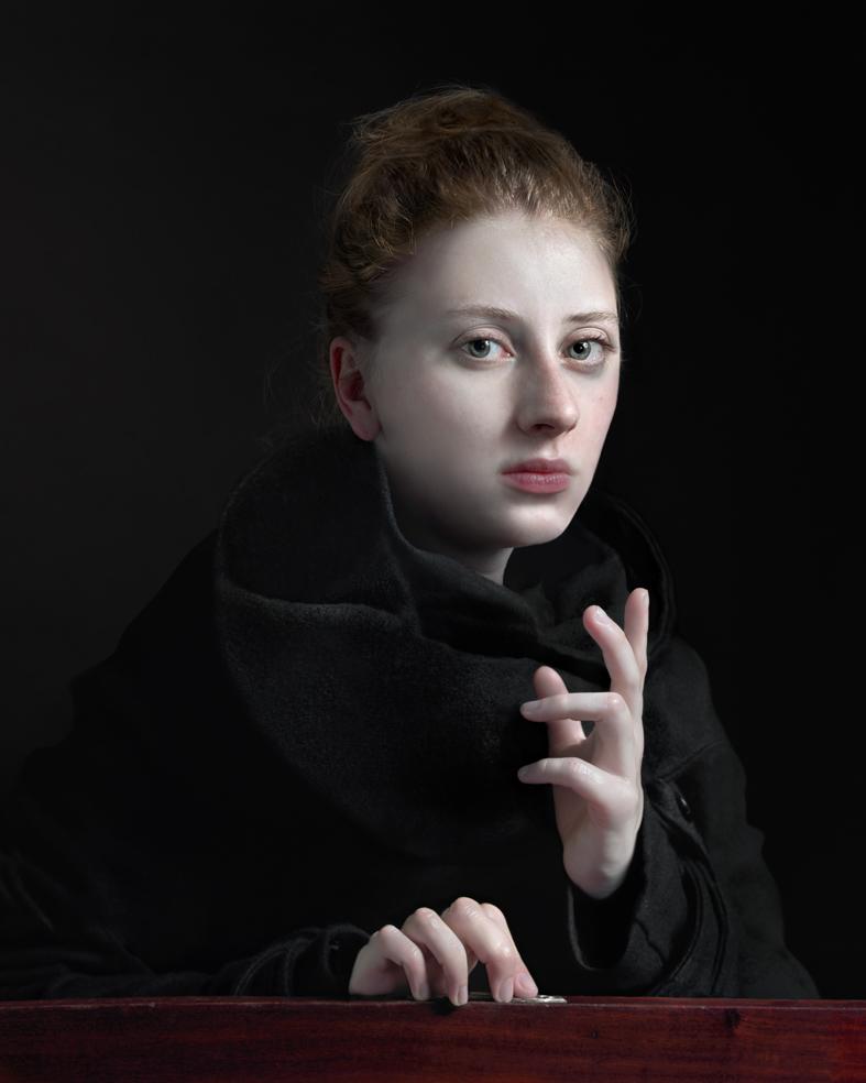 A young woman with reddish-brown hair styled upward gazes toward the camera against a dark background. She wears a black garment and rests her pale hands on a dark wooden surface, her expression contemplative and direct.