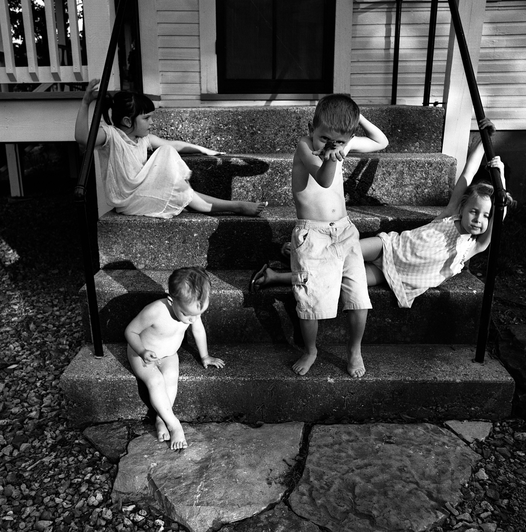 Black and white photograph of five young children positioned on concrete steps outside a residential building. The children, ranging from toddlers to early elementary age, are arranged across multiple step levels in various playful poses. The composition captures a candid domestic moment with natural architectural framing provided by the stepped structure and the house's siding visible in the background.