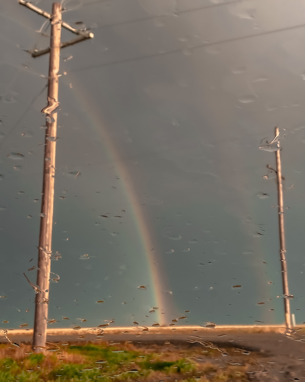 Two weathered wooden poles stand in a flooded landscape beneath a stormy gray sky, with a luminous rainbow arc stretching between them. The scene captures an ethereal interplay of light and water, with scattered debris visible on the wet ground.
