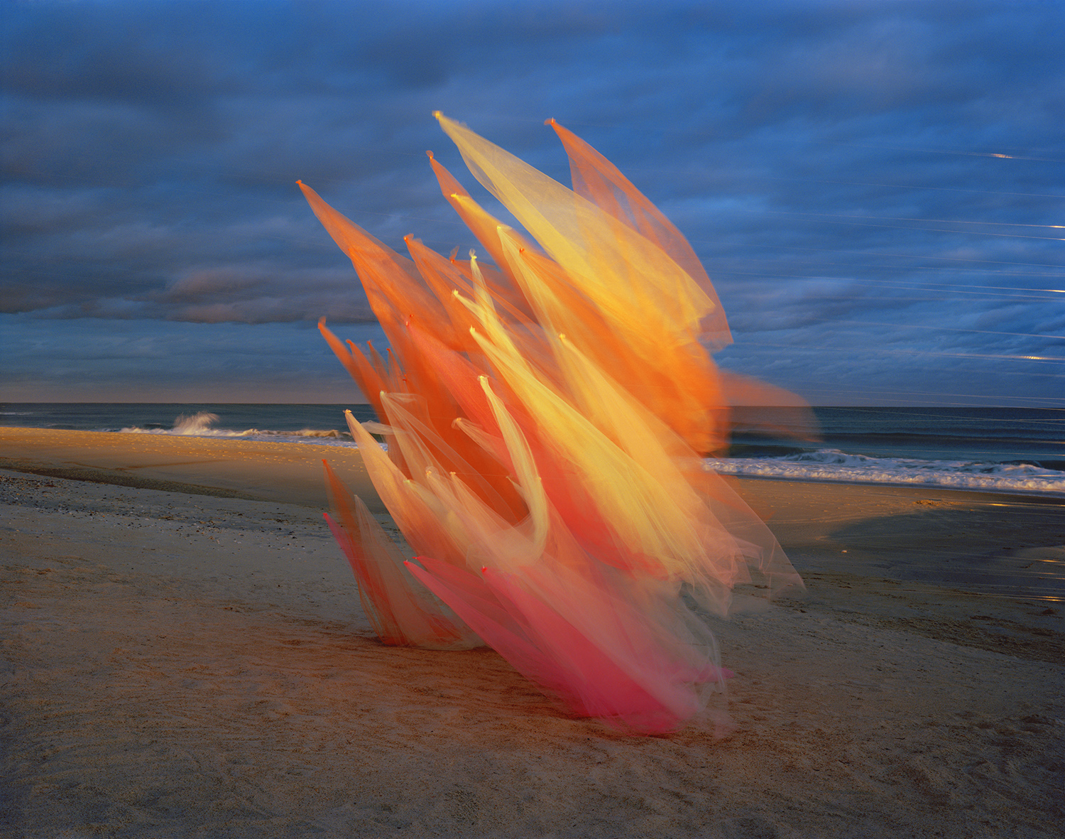 Translucent fabric in graduated shades of coral, orange, and yellow billows on a beach at dusk, creating sharp angular peaks against a blue-grey sky and ocean horizon.
