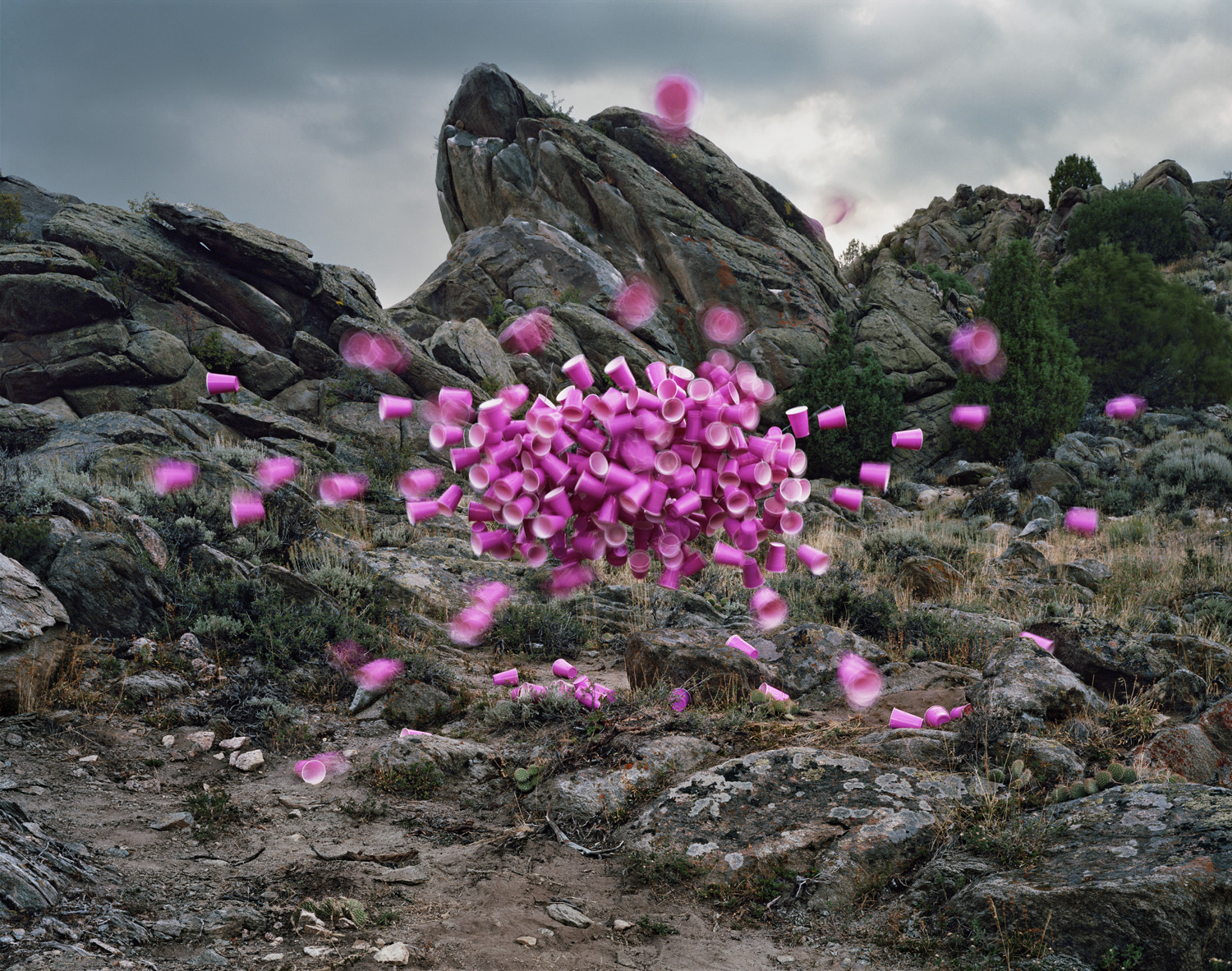 Vibrant pink plastic cups are suspended and scattered in mid-air across a rugged desert landscape dotted with large boulders and sparse vegetation, creating a surreal contrast between the artificial, everyday objects and the natural geological formations.