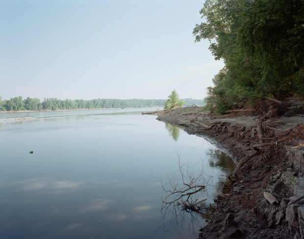 A calm river stretches across the composition, its mirror-like surface reflecting overcast sky and distant tree-lined banks. In the foreground, an eroded earthen bank slopes steeply toward the water's edge, with exposed soil and sparse vegetation, while a small dead branch protrudes from the bank into the shallow water.