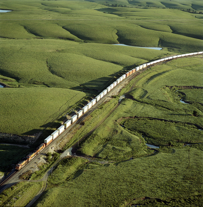 Aerial view of a long freight train with white and colored cars winding through rolling green prairie hills, casting shadows across the undulating landscape with patches of water visible in the distance.