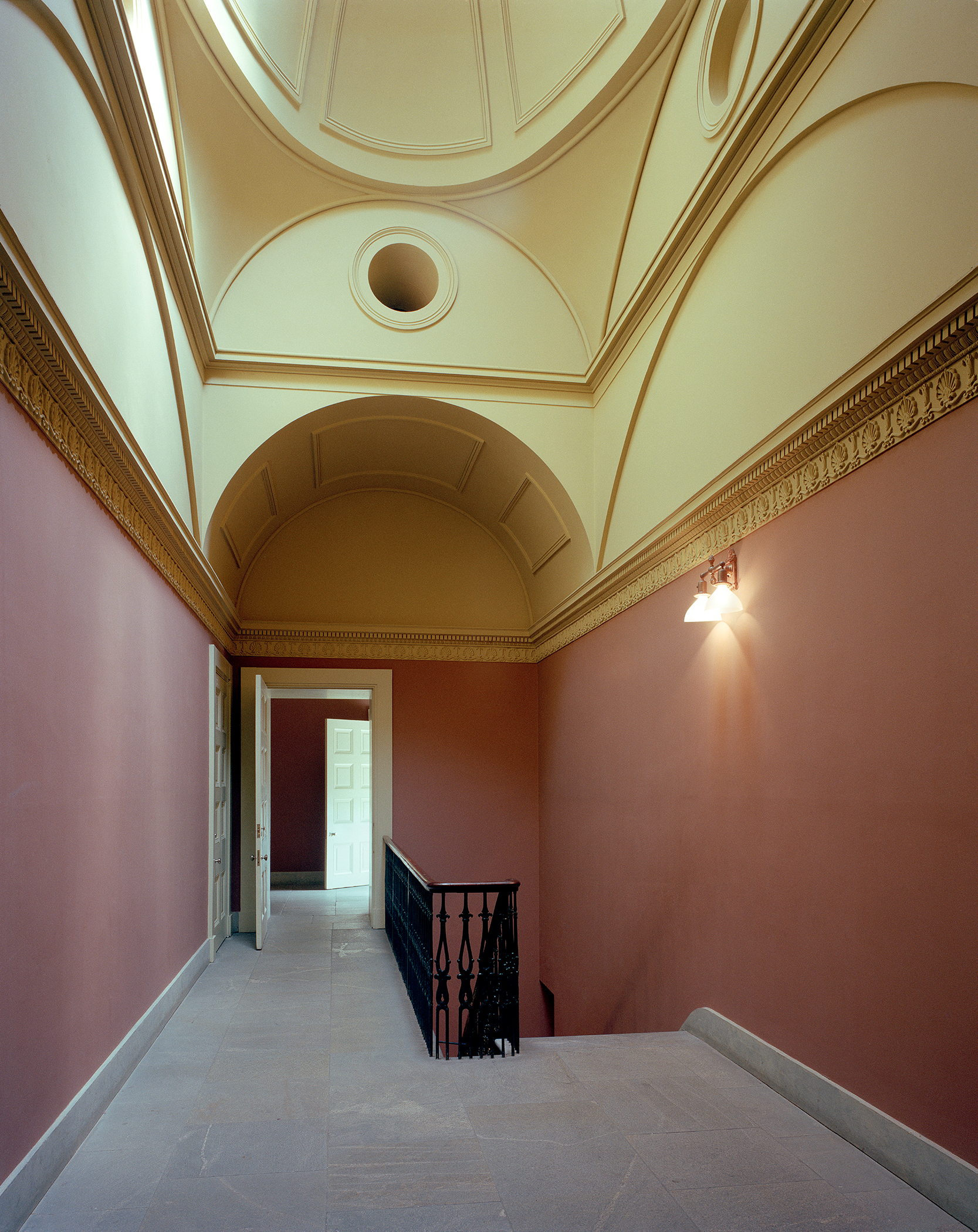 A classical architectural corridor with terracotta-red walls and cream-colored ceiling features ornate molding and a barrel vault. A black wrought-iron railing frames an opening on the right, while a doorway with white trim leads deeper into the space ahead, creating a sense of perspective and geometric symmetry.
