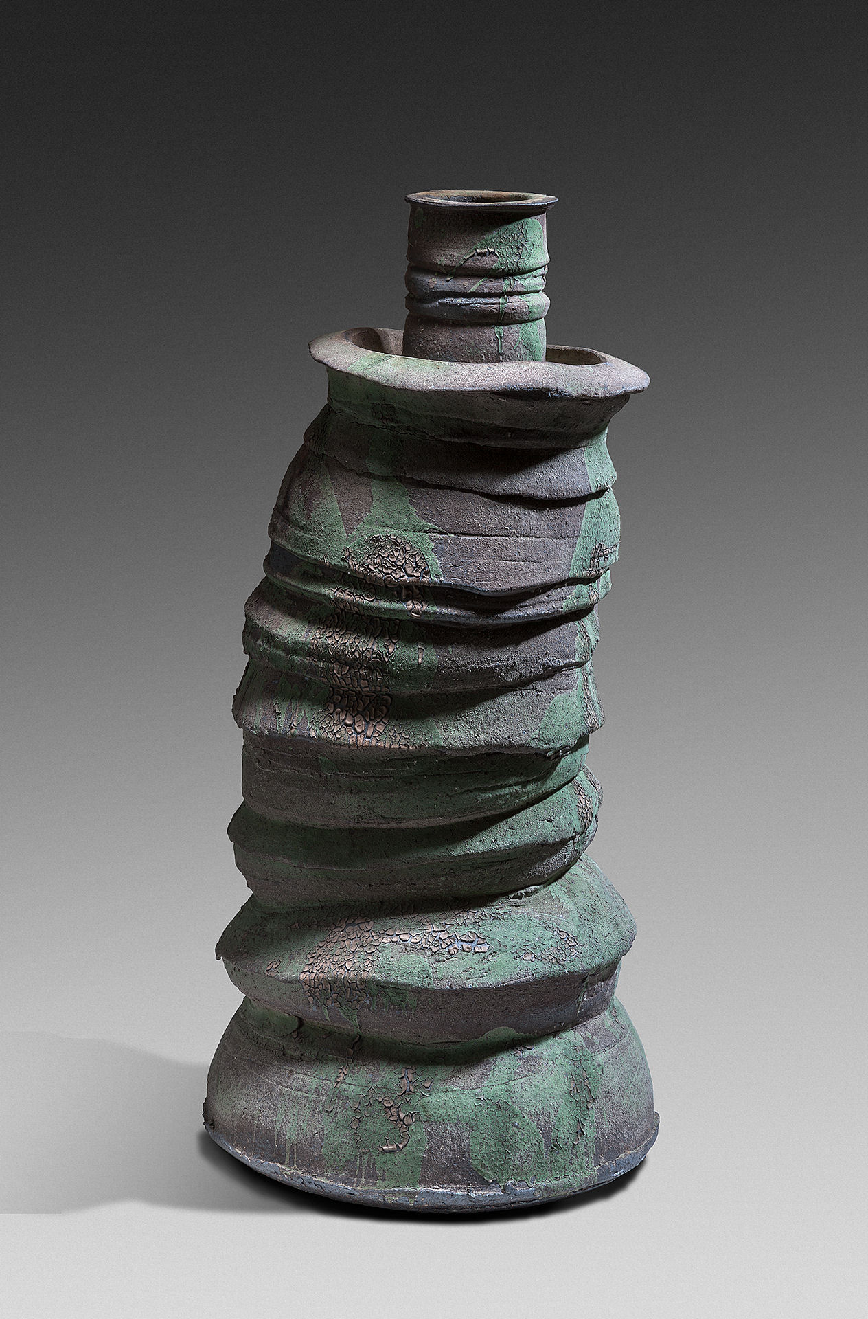 Tall cylindrical ceramic vessel with a conical base, composed of stacked horizontal bands that create a rippled, organic form. The dark stoneware surface is covered with green and gray patina, suggesting oxidation or intentional surface treatment.