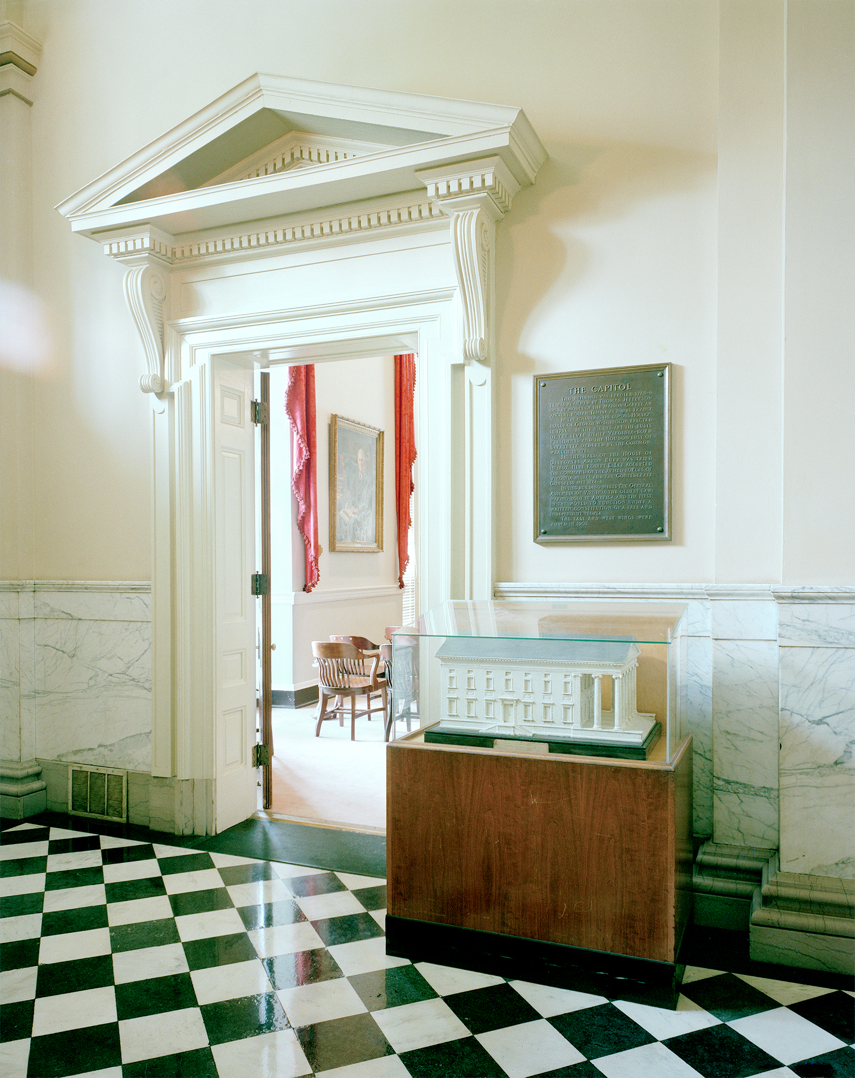 Interior view of a neoclassical Capitol building entrance featuring a grand white doorway with decorative pediment and fluted columns. A wooden display case with a glass top sits in the foreground on a green and white checkered marble floor, with a commemorative plaque mounted on the wall above. The space beyond shows red drapery and period furnishings visible through the open doorway.