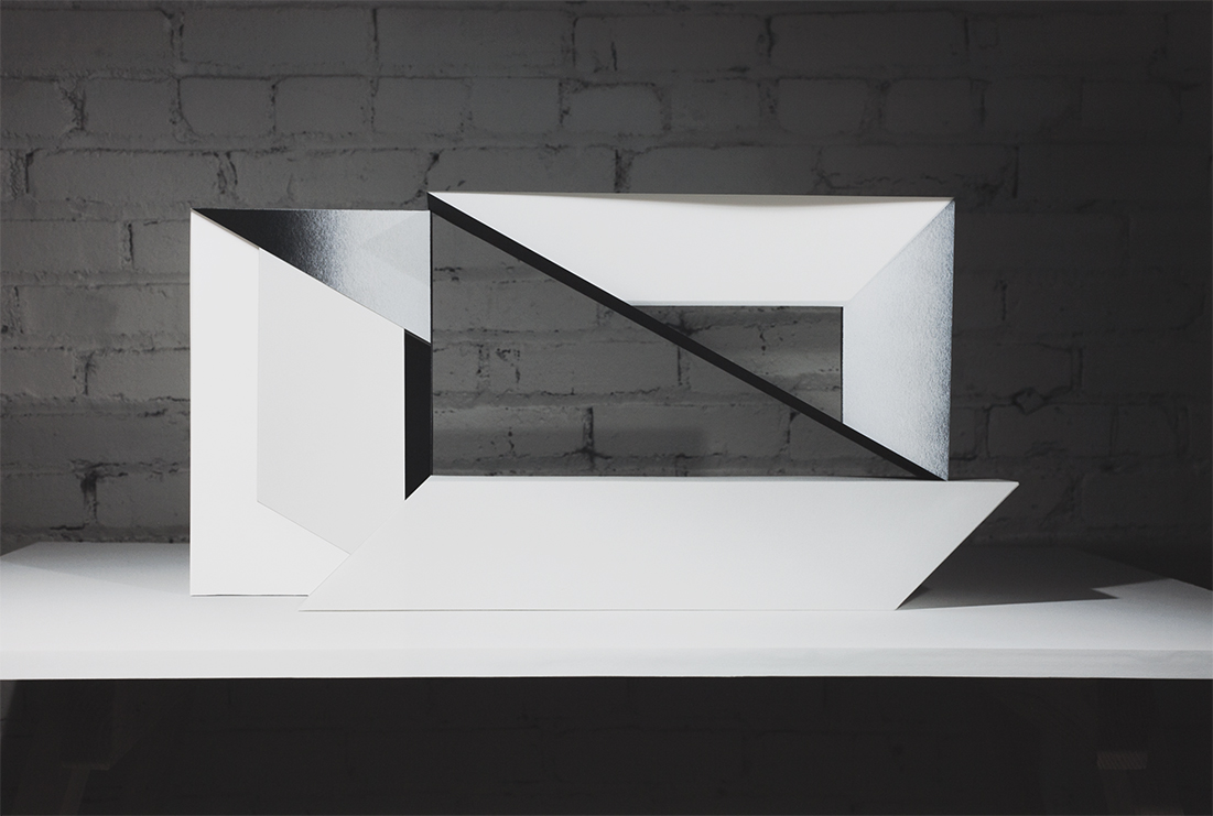 A white geometric paper sculpture with sharp angular cuts and folds sits against a dark brick wall. Black diagonal lines and negative spaces create visual depth within the layered, three-dimensional form, contrasting with the white paper surface.