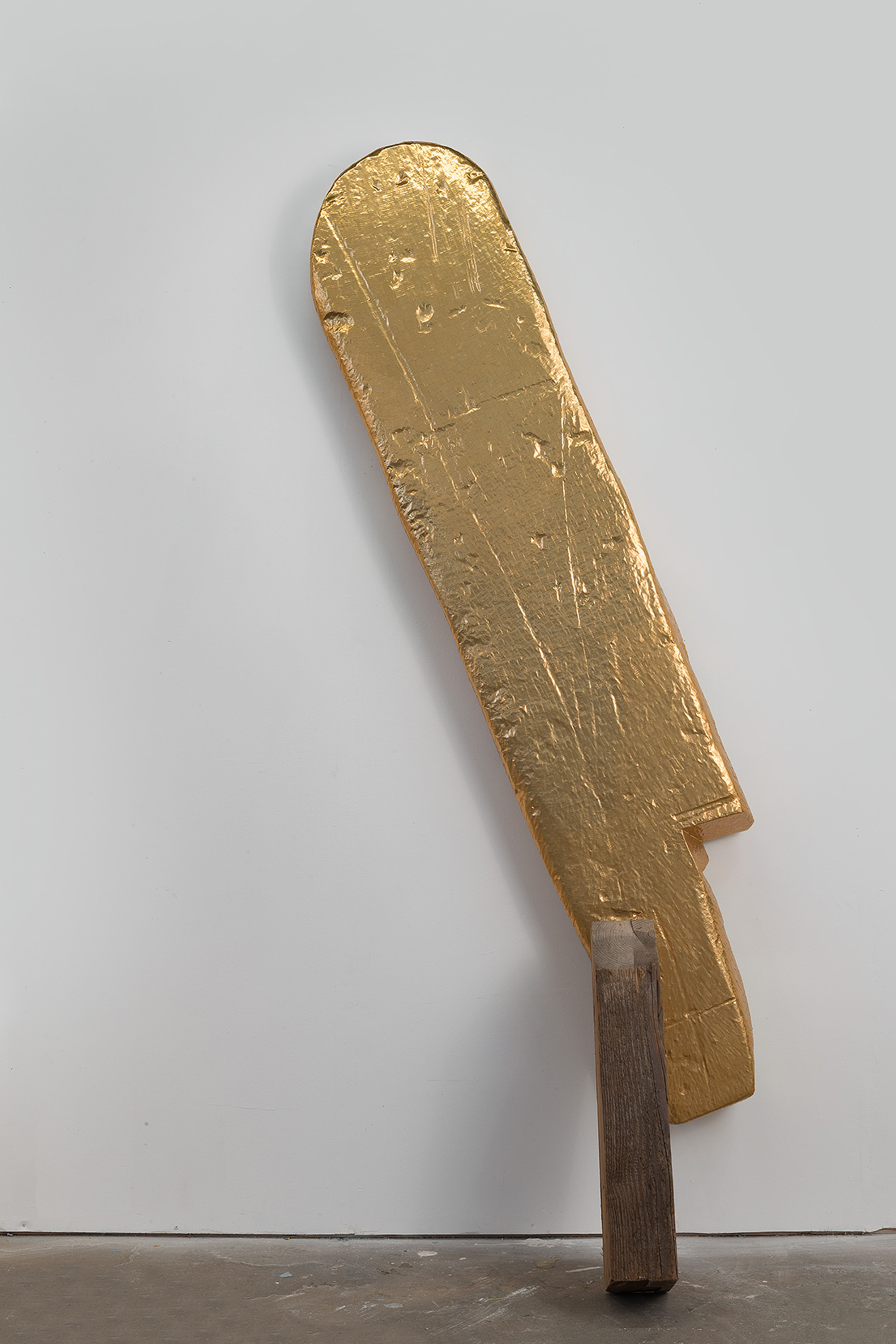 A tall, vertical sculptural form with a rounded top rendered in gold leaf mounted on a dark wooden post or beam. The surface is heavily textured with scratches and weathering, creating an abstract, weathered appearance against a white gallery wall.