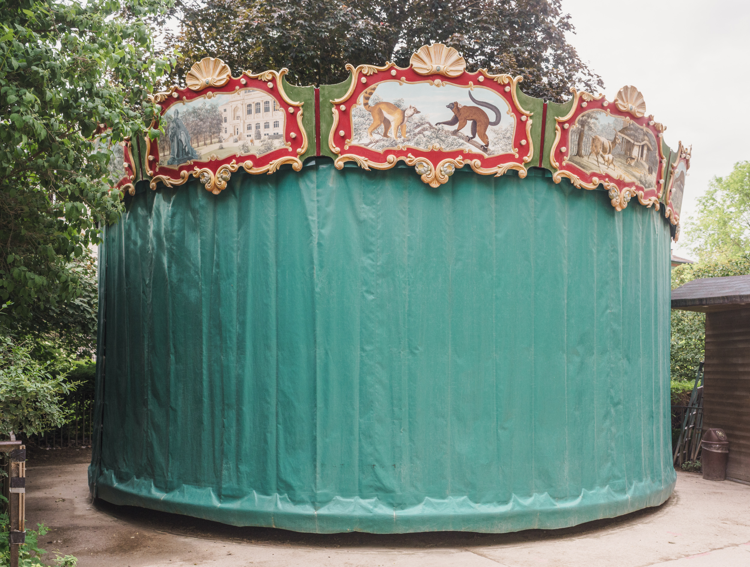 A large cylindrical structure with oxidized copper-green patina is topped with an ornate red and gold decorative carousel-style crown featuring shell motifs and painted panels depicting monkeys and architectural scenes. The structure stands in a wooded outdoor setting.