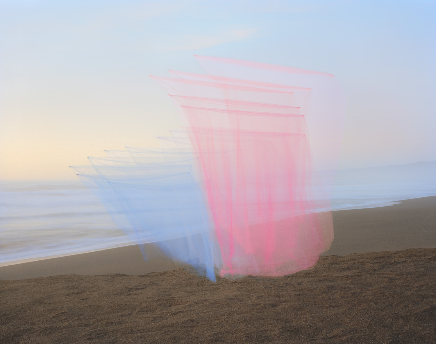 Translucent layers of pink and blue tulle fabric billow on a sandy beach at dusk, creating ghostly, overlapping rectangular forms against the soft horizon and calm ocean waters.
