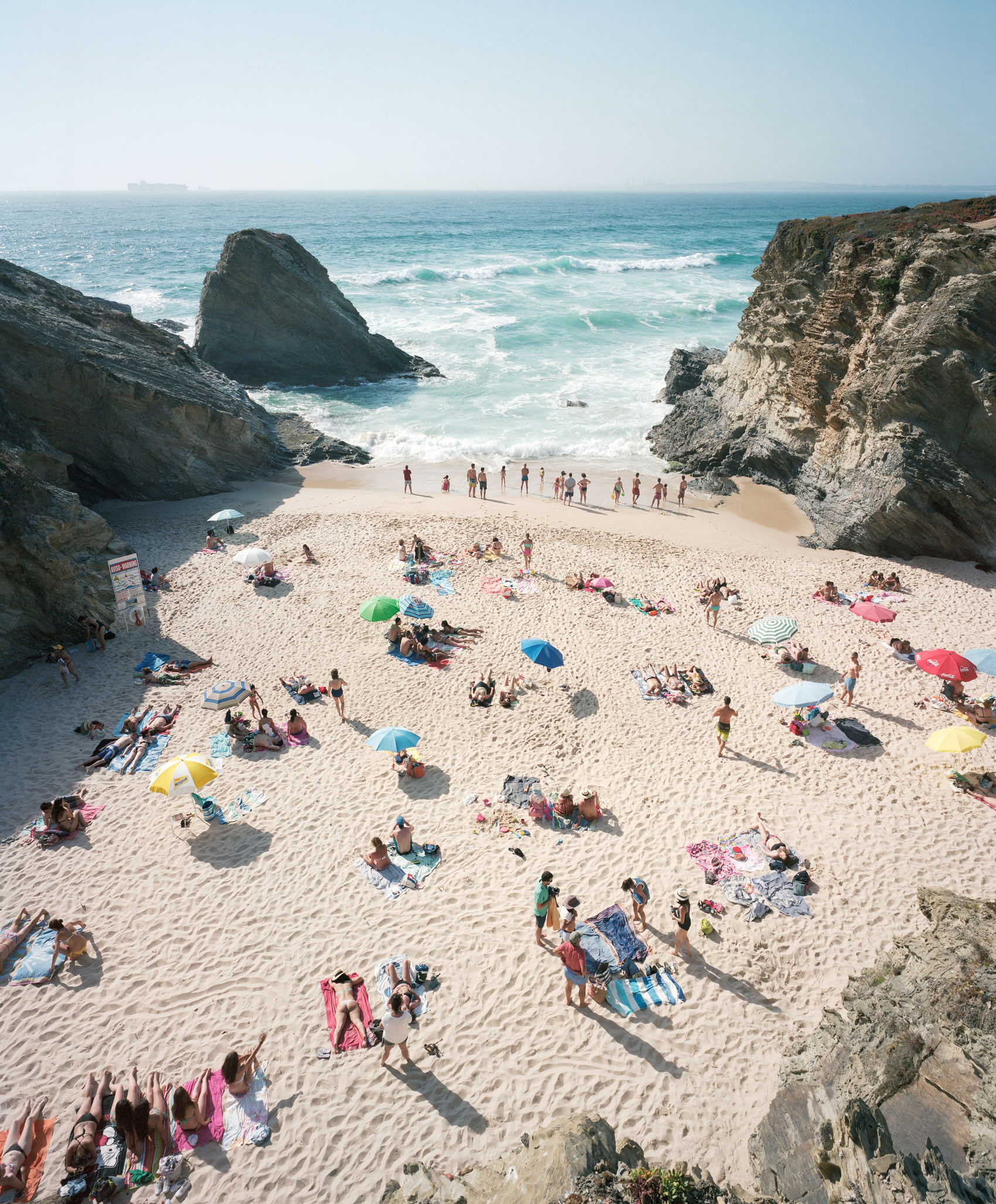 Aerial view of a crowded beach cove nestled between dramatic rocky cliffs, with numerous beachgoers scattered across the sand under colorful umbrellas in shades of blue, pink, yellow, and green, while turquoise waves break against the shore and a distant ship appears on the horizon.