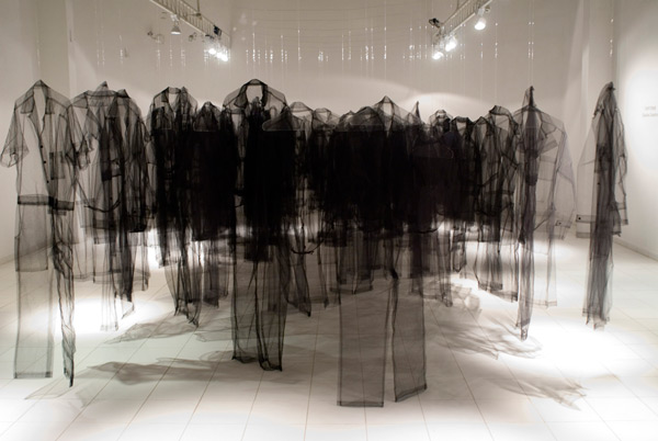A large-scale room installation featuring numerous translucent black tulle garments suspended from above, creating silhouettes of human figures that overlap and layer throughout the white gallery space, with dramatic shadows cast by overhead lighting.