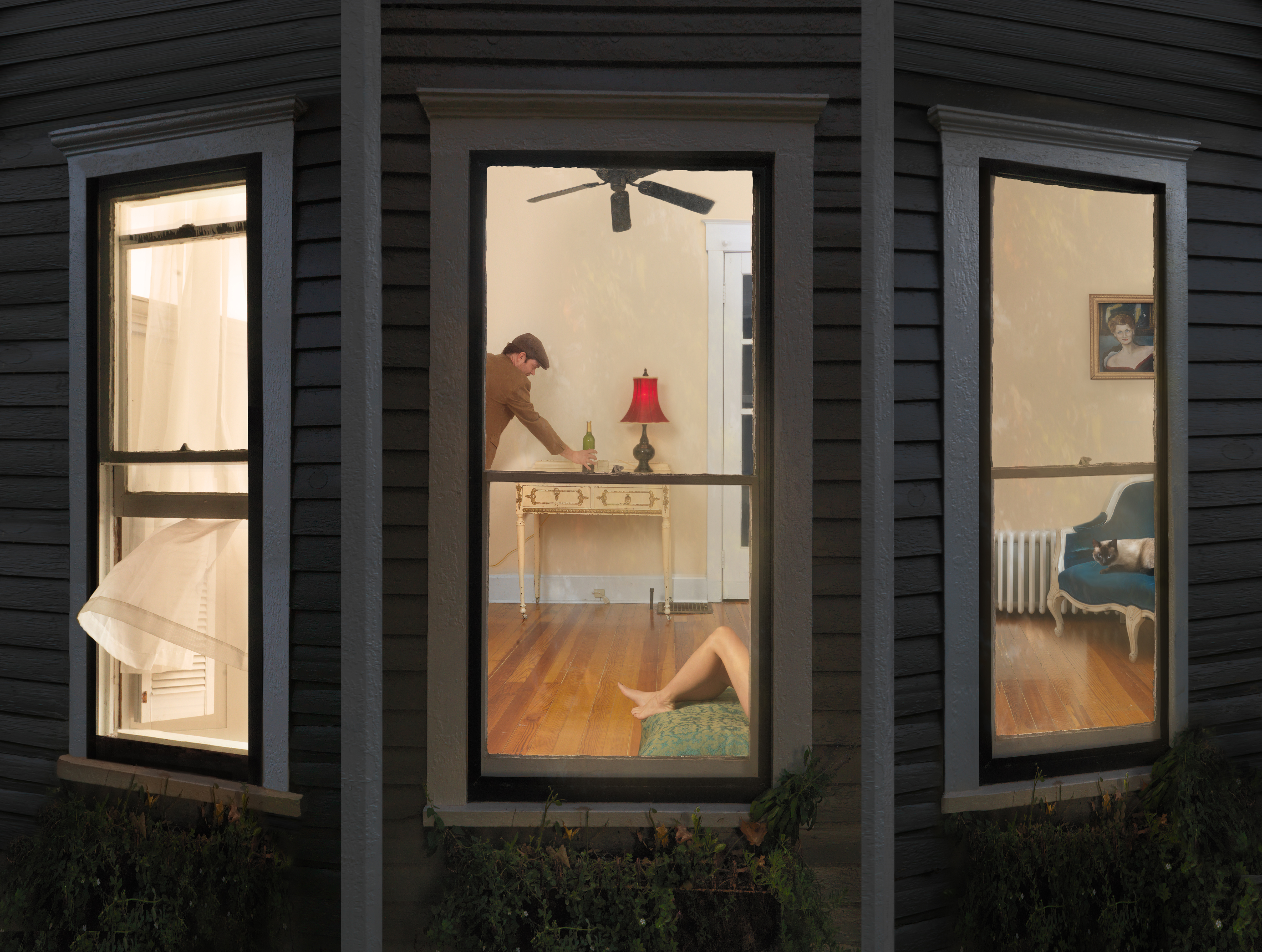 Viewed through three illuminated windows of a dark wooden house at night, an interior domestic scene shows a woman in a green skirt reclining on a wooden floor in the foreground, while a man in a brown shirt leans over a table with a red-shaded lamp in the background. The warm, staged lighting and composed arrangement of furniture, including a blue chair and framed portrait on the wall, creates a theatrical narrative across the sequential window frames.
