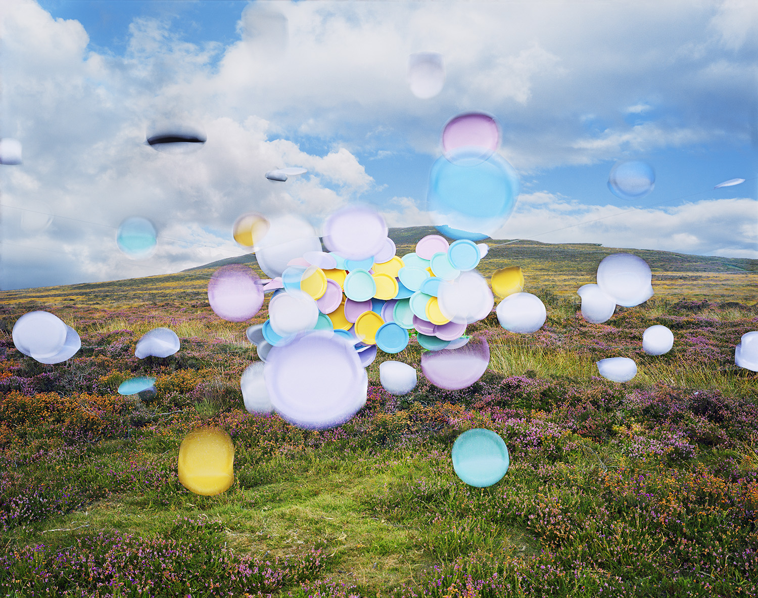 Colorful translucent spheres in white, pink, blue, and yellow float above a moorland landscape dotted with purple heather and green grass, with rolling hills visible under a cloudy sky.