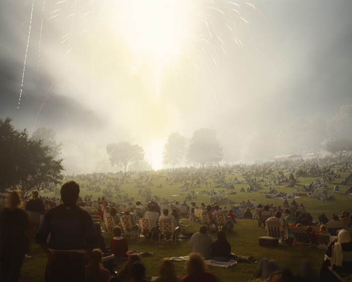 A large crowd of silhouetted figures scattered across an expansive green field beneath a hazy, luminous sky with fireworks bursting above. Atmospheric mist and golden light create a dreamy, ethereal quality over the gathered masses.