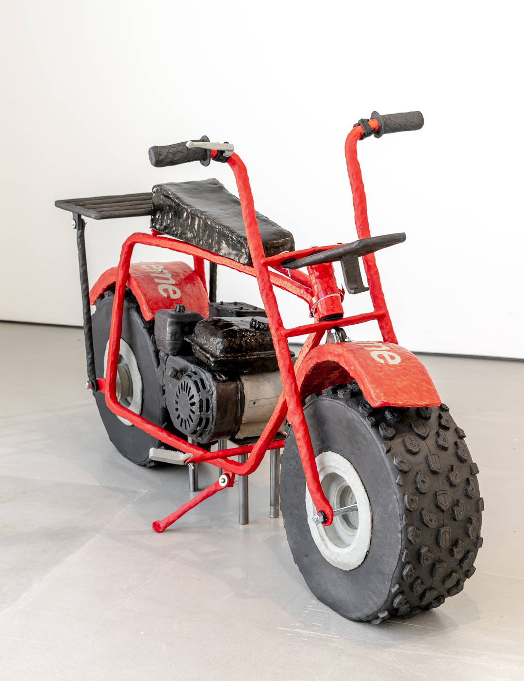 A red and black sculptural motorcycle with an oversized front tire, featuring handlebars, a seat, and various mechanical details rendered in ceramic and steel. The work sits on a gallery floor against a white wall, presented as a functional-looking yet deliberately exaggerated miniature vehicle.