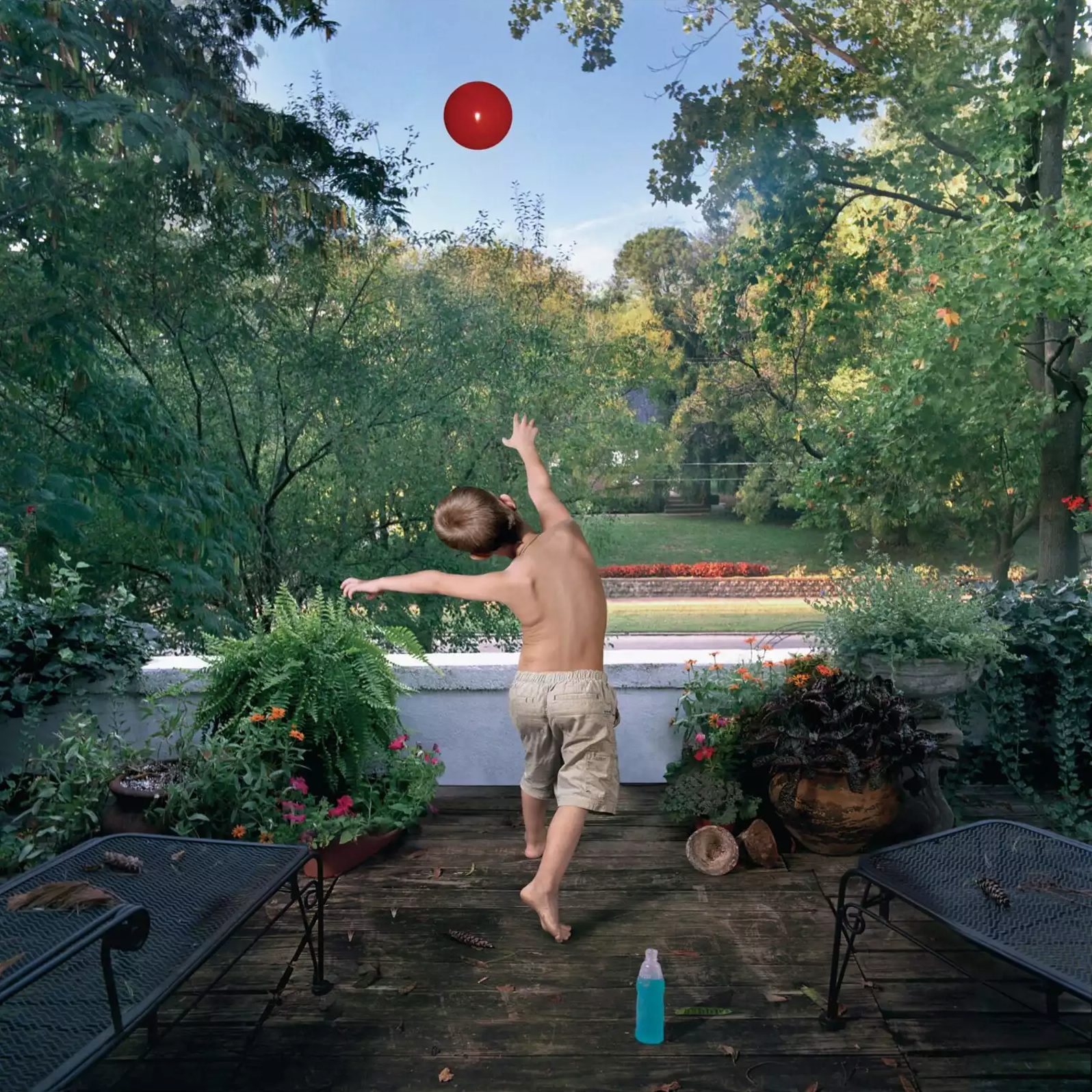 A shirtless child jumps on a wooden deck, reaching toward a bright red ball floating in the sky above a lush garden. Surrounded by flowering plants, lounge chairs, and trees, the scene captures a moment of gleeful play in an idyllic outdoor setting.