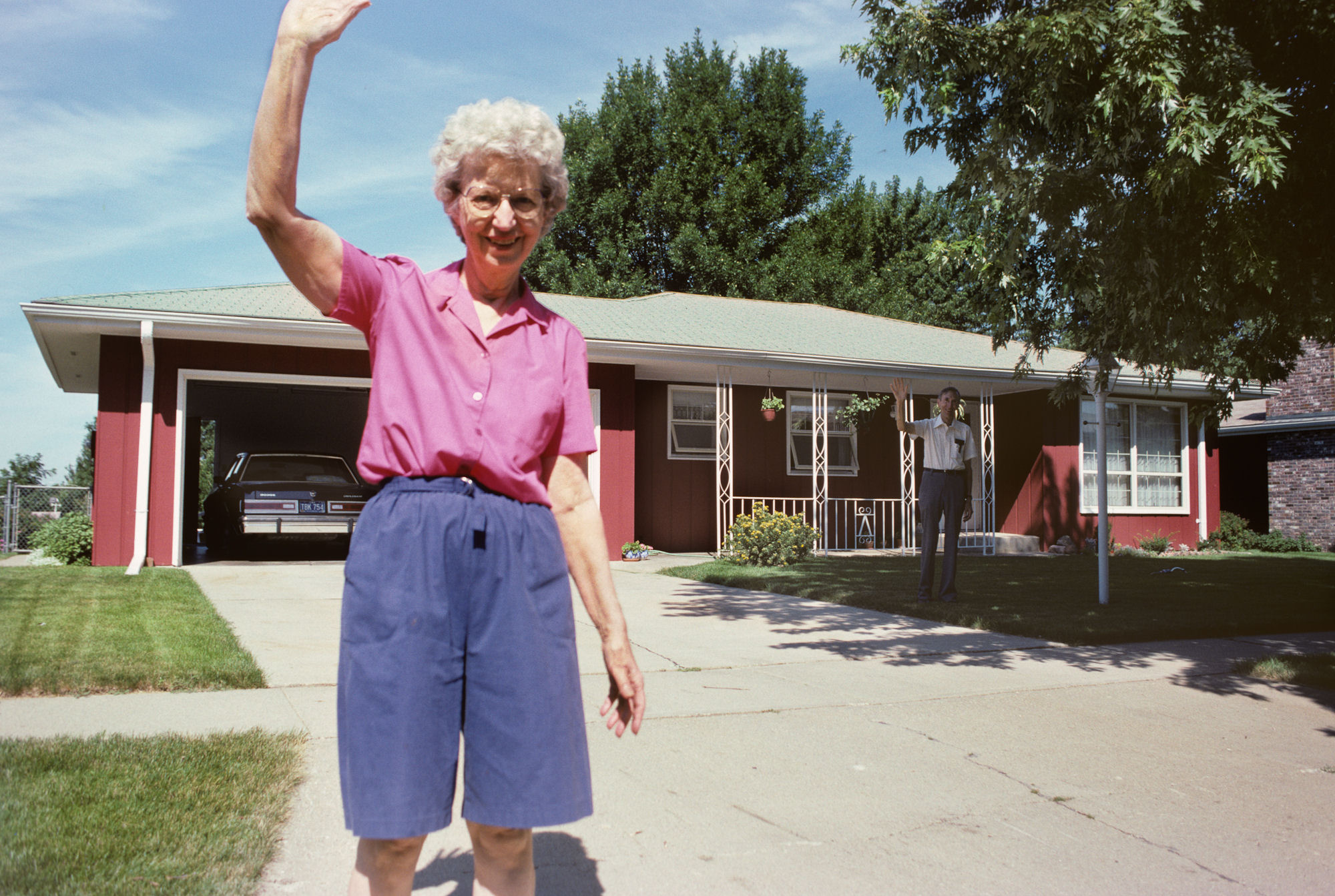 An elderly woman with white hair wearing a pink short-sleeved shirt and blue shorts stands in a residential driveway, waving with her raised arm. Behind her is a mid-century modern ranch-style house with red and brown exterior walls, a carport with a car visible inside, and manicured lawn. A second figure can be seen standing on the house's porch. The photograph has the warm color palette and composition style of 1990s color film photography.