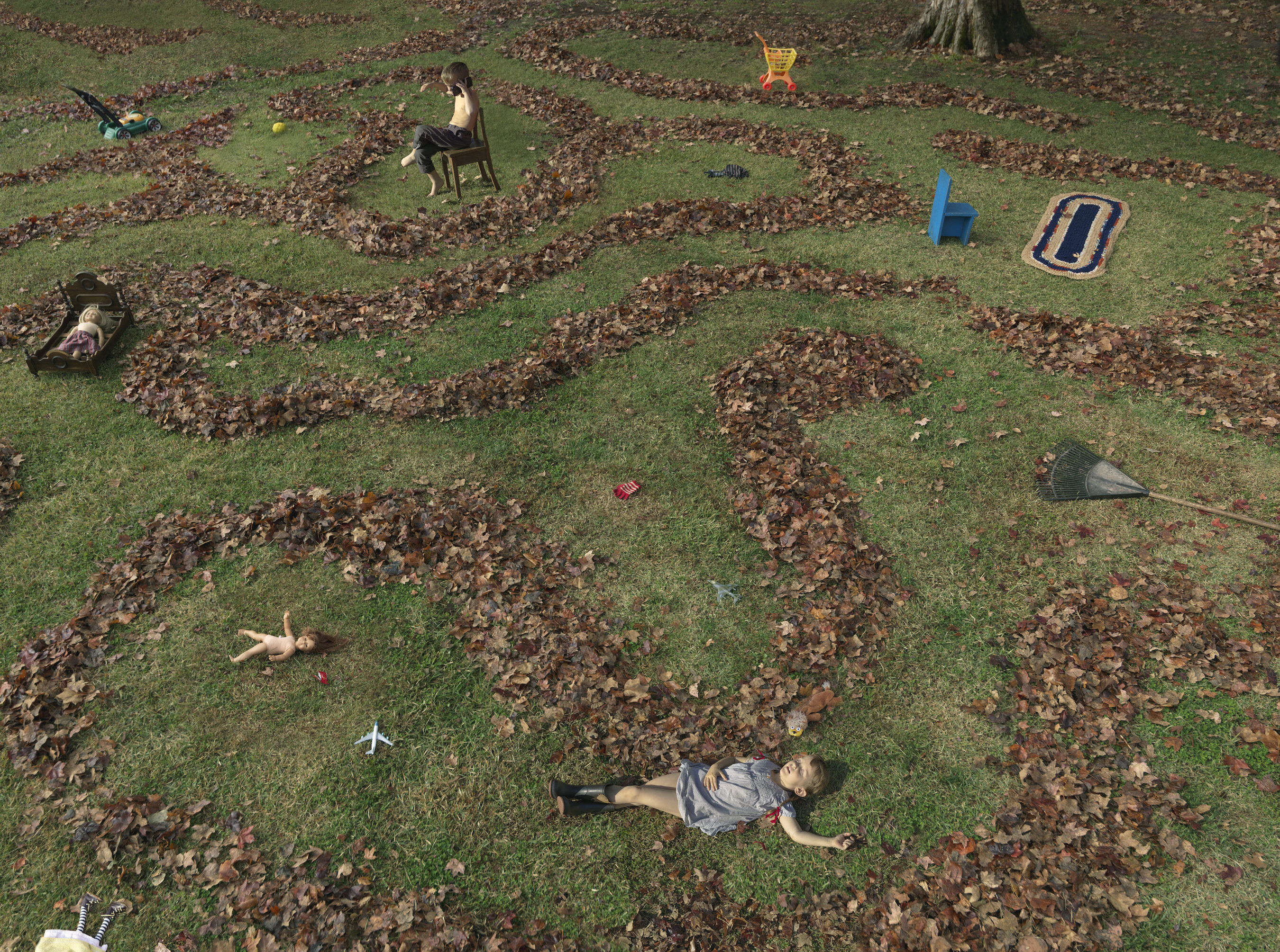 Aerial view of a grassy yard scattered with brown fallen leaves arranged in organic patterns, with two children lying on the grass among the leaves and various toys and objects dispersed throughout the scene, including a chair, rake, and colored play items.