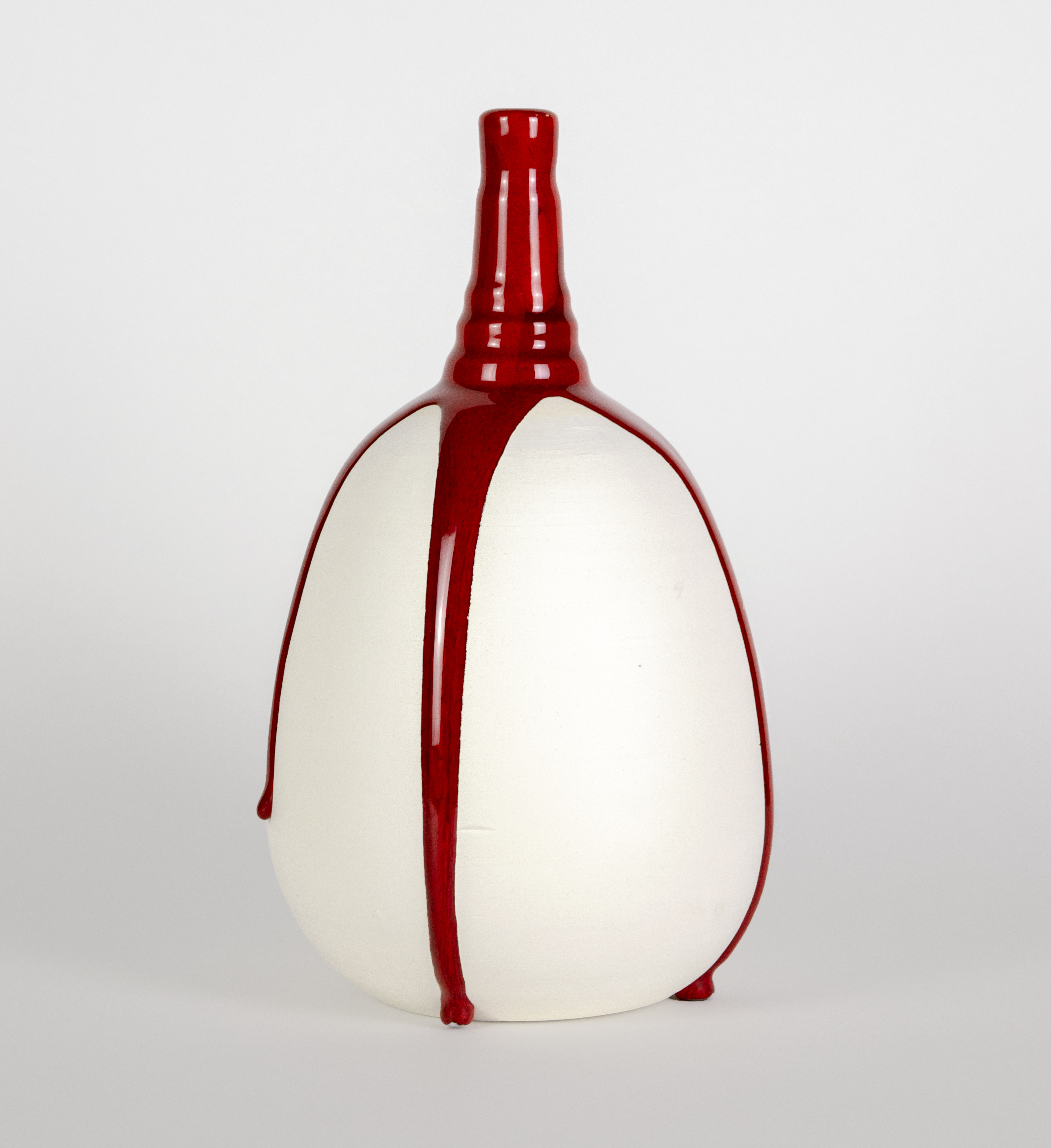 A rounded, cream-colored vessel with a tapered red neck and spout, bound vertically by bold red straps that extend from top to bottom, creating a striking contrast between the soft neutral body and vibrant crimson accents.