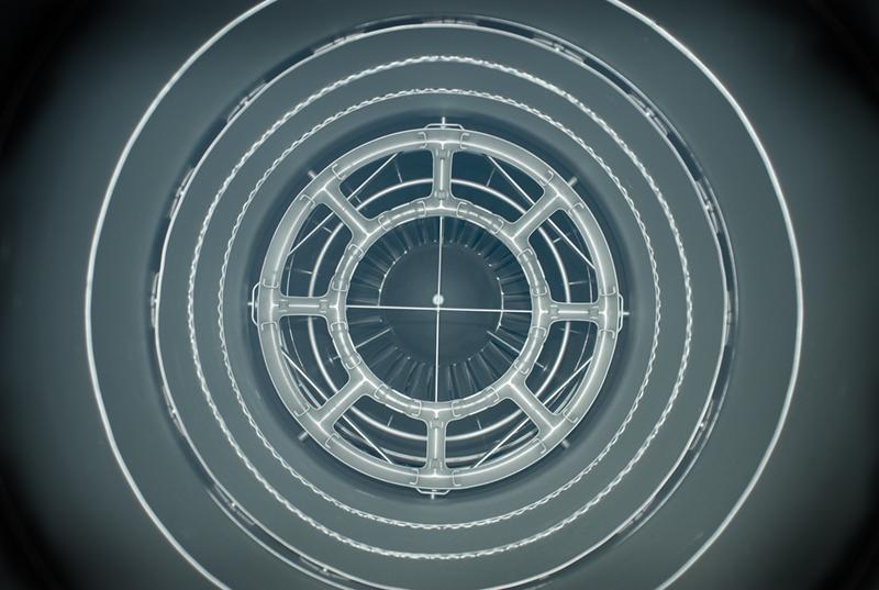 Concentric rings of translucent geometric forms radiate from a central circular structure, creating a tunnel-like perspective. The composition features repeating patterns of architectural or mechanical elements rendered in pale gray and white tones against a darker background, suggesting depth and symmetry.